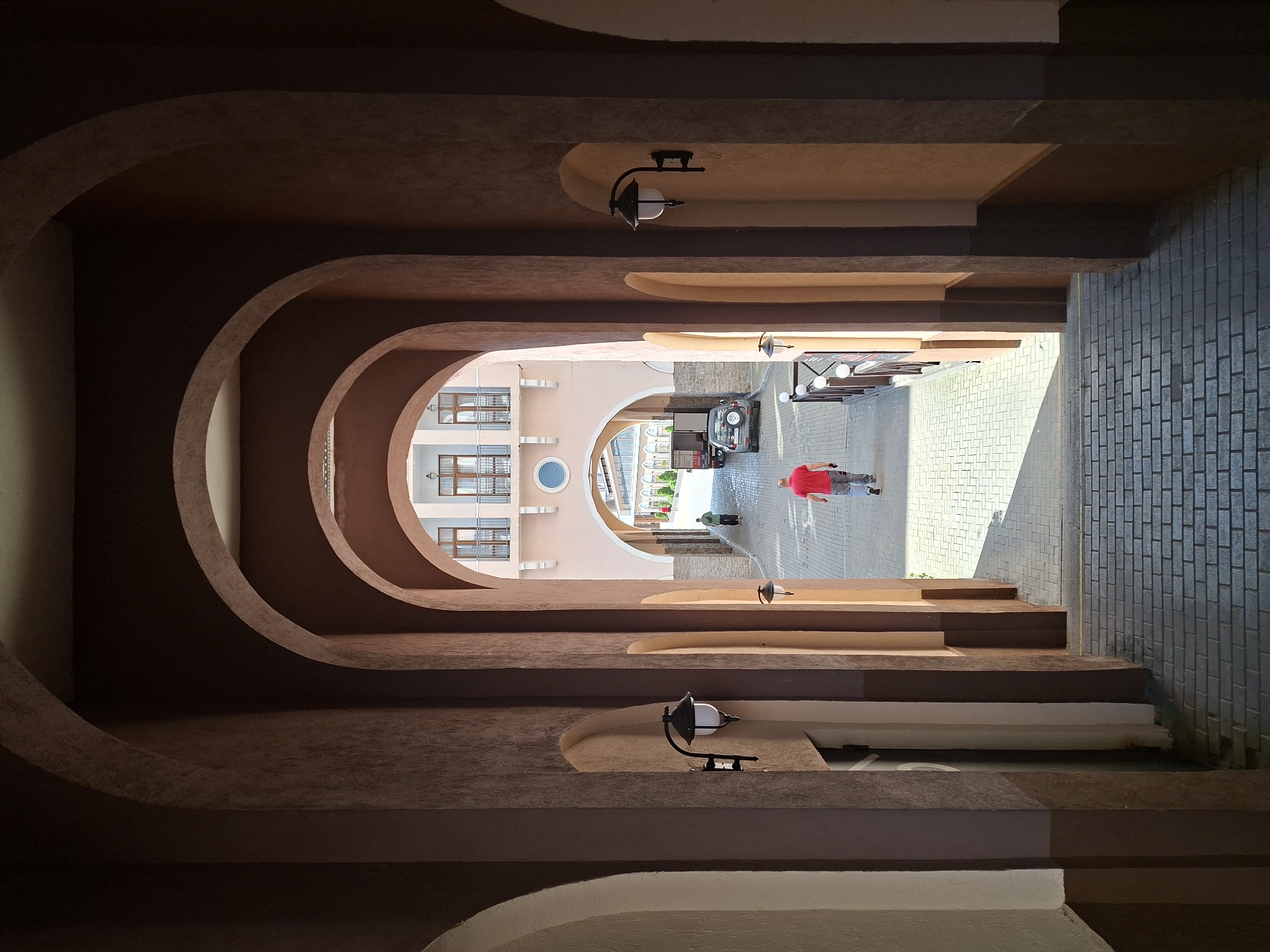 An outdoor view through a series of arched passages in Estosadok, Russia. The perspective is looking down a tiled walkway, framed by dark brown and tan arched pillars. On the left side, a white door is visible. On the right, a black outdoor lamp is mounted on a pillar.
Further down the walkway, sunlight casts shadows on the patterned tiles. A man wearing a red shirt and gray pants walks away from the camera, towards a pink building at the end of the passage. To his left, another person in a green shirt walks away. A dark gray SUV is parked on the right side of the walkway. The pink building has multiple windows with balconies and a circular window.