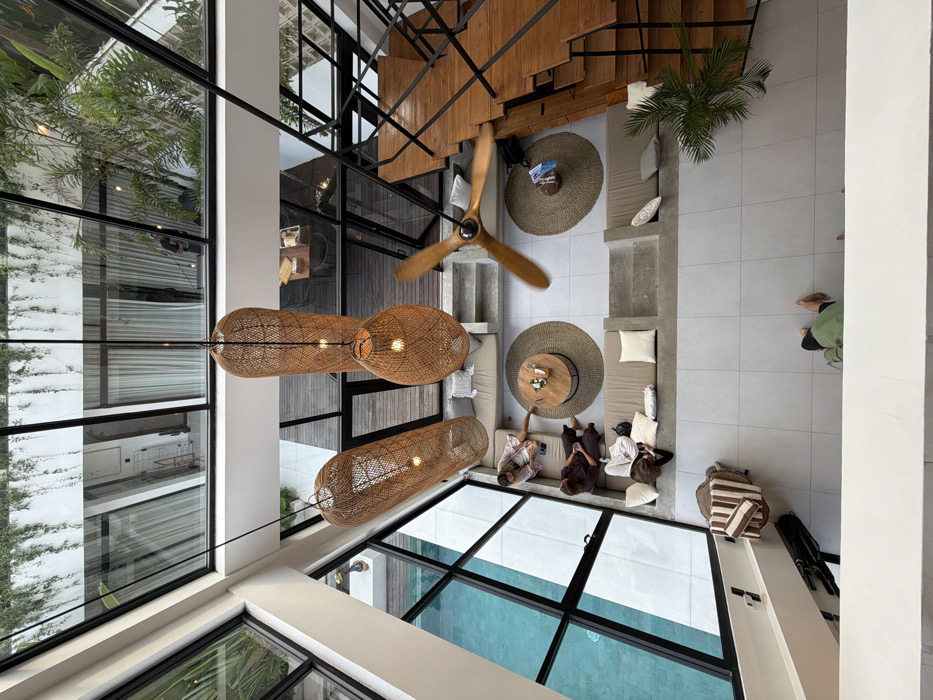 The image presents an elevated, downward-looking view into a modern, multi-level architectural interior, identifiable as a villa or communal space in Canggu, Indonesia. The central area features a light-tiled floor and a high glass ceiling structure with visible greenery above. Three large, illuminated pendant lights, constructed from woven rattan in varying elongated and rounded shapes, hang from the ceiling. A three-bladed wooden ceiling fan, blurred due to motion, is positioned to the right of the central lights.
On the left, expansive glass panels form a wall, providing a clear view of an outdoor swimming pool with bright blue water. Adjacent to this wall, a built-in lounge area features light-colored cushions on concrete bases. Three individuals are seated here: two women on the left-most sofa, one in a light robe and the other in a darker garment, both looking downwards. A circular woven rug anchors a round wooden coffee table holding small items. A third person sits on a lower-level sofa, also engaged with a mobile device. A fourth individual is partially obscured on a sofa further to the right.
To the right of the scene, a substantial wooden staircase with black metal railings ascends. Near the staircase, a second circular woven rug and a smaller round wooden table are visible. A small potted palm plant sits on the floor nearby. In the lower-left foreground, a striped canvas bag and a dark backpack rest on the tiled floor. At the very bottom edge of the frame, the bare feet and lower torso of a person wearing a green top and dark bottoms are visible. Reflections in the overhead glass reveal parts of the building's exterior and the figure of a person, possibly a male wearing a dark shirt, standing outside the structure.
