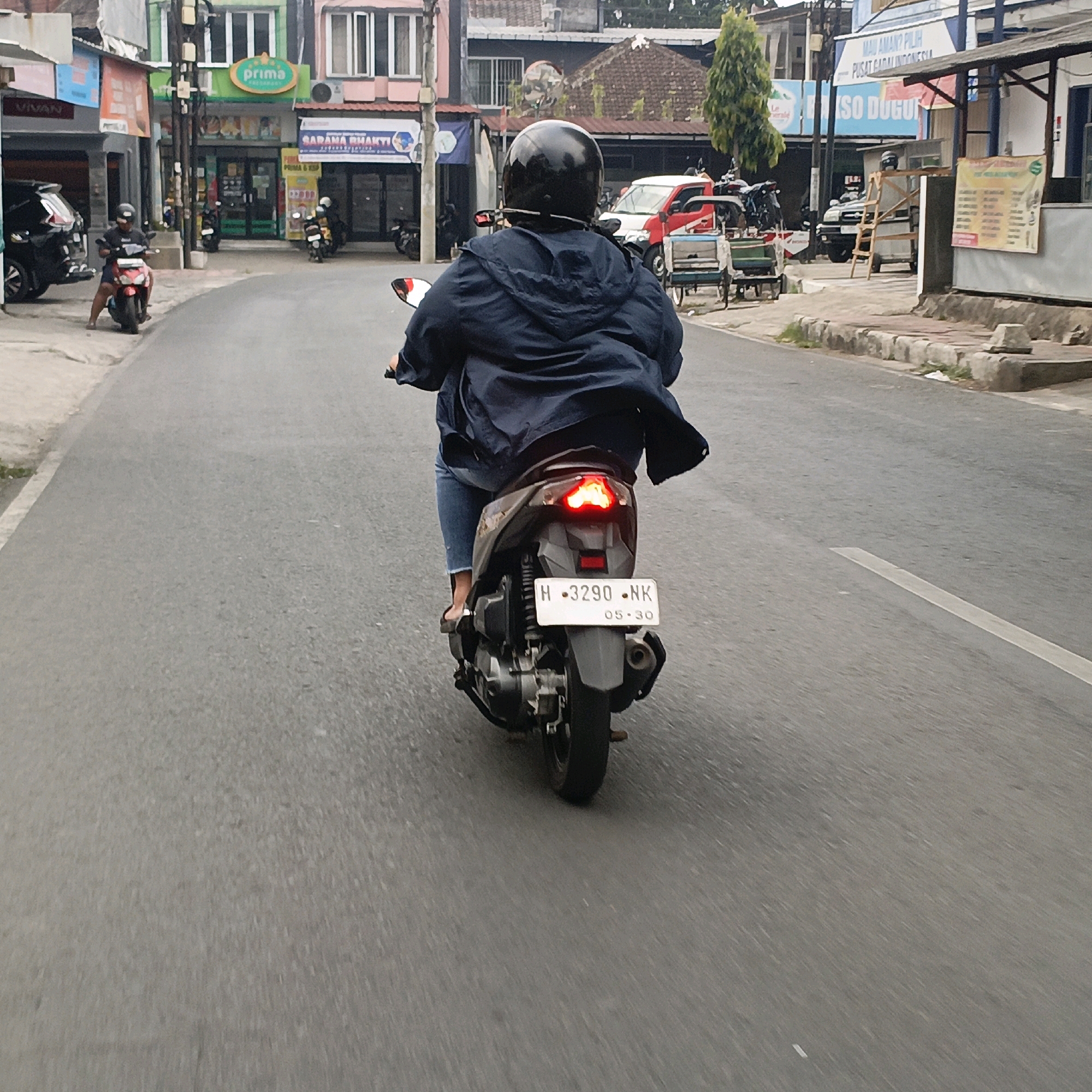 The image captures a street scene in Salatiga, Indonesia, from the perspective of someone riding on a motorcycle. In the foreground, the rear of a motorcycle is visible, with the rider wearing a dark blue jacket and jeans. The motorcycle's license plate reads "H · 3290 · NK" and "05·30". The brake lights of the motorcycle are illuminated, indicating it is either braking or has just braked. The road is paved and appears to be a street in an urban or semi-urban area, with a white line marking a lane on the right side.
The background reveals a row of buildings lining the street, suggesting a commercial area. One building, painted green and white, has a sign that reads "prima". Another building has a sign with text that includes "SARANA BHAKTI". Further down the street, there are more buildings and structures, including some with signs for food establishments such as "BASO DUGUR". Various vehicles, including other motorcycles and a red car, are parked or in motion on the street and sidewalk. There are also some carts or stands on the side of the road. The overall environment suggests a typical day in a bustling Indonesian city.