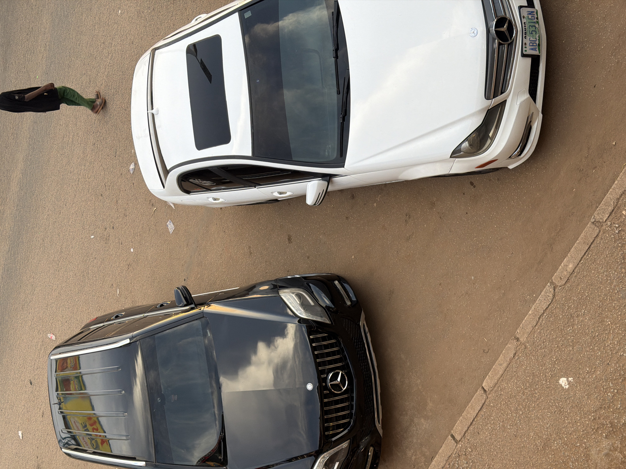Two Mercedes-Benz cars, a black SUV and a white sedan, are parked on a dusty, unpaved road in Bukuru, Nigeria. The black SUV, positioned on the left, is a larger model, possibly an ML-Class, with its front grille and headlights visible. The white sedan, on the right, is a C-Class model, with its license plate reading "ABC 531GN". A person, wearing a black robe-like garment and green trousers, walks away from the cars in the background. The ground is covered in reddish-brown dirt, with scattered white debris. A concrete curb separates a more heavily paved area on the left from the dirt road. The scene appears to be outdoors, likely in a residential or commercial area with minimal infrastructure.