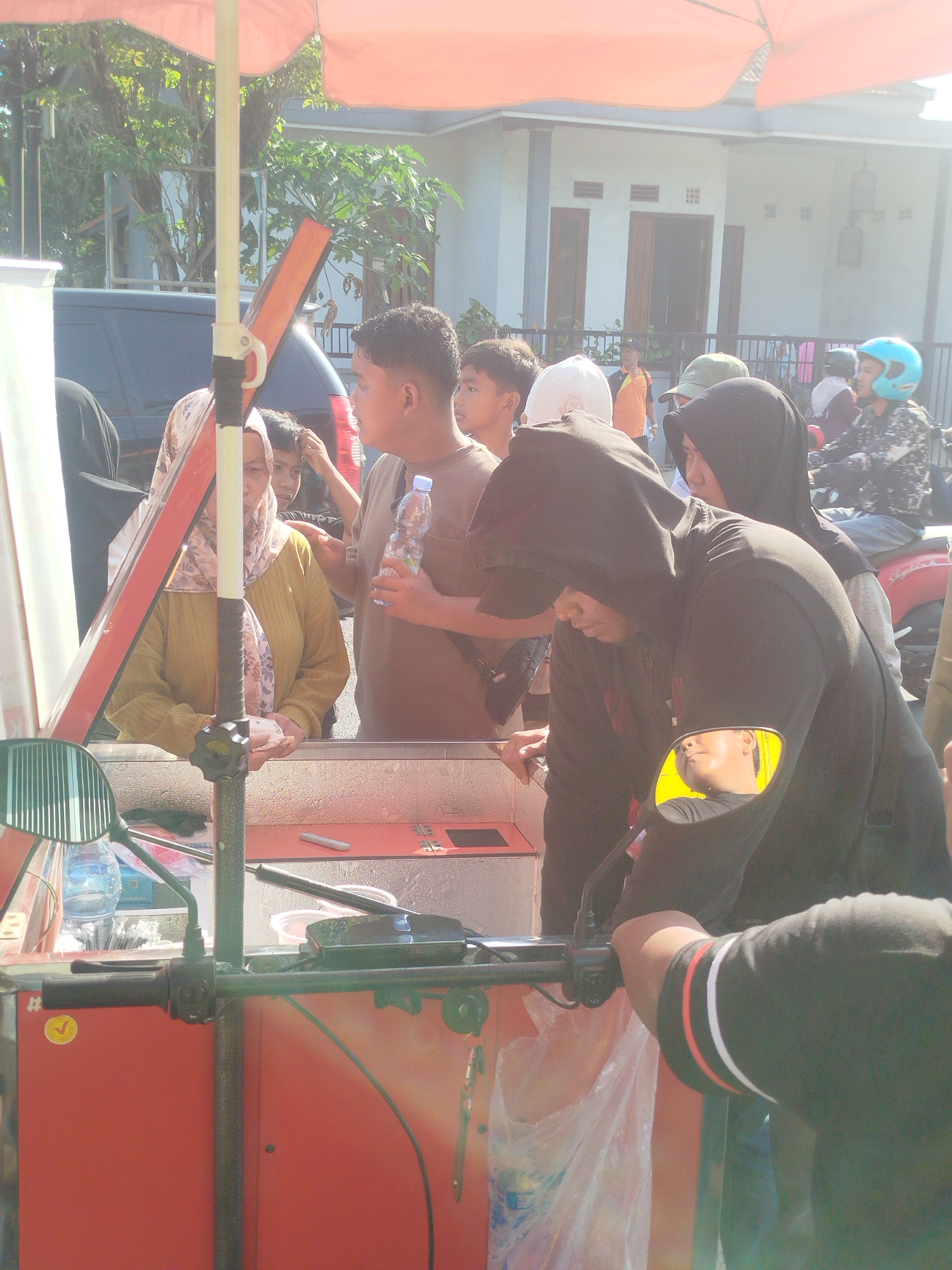 An outdoor scene in Burneh, Indonesia, shows a group of people gathered around a food cart under a large orange umbrella. A man in a brown shirt holds a bottle of water, speaking to a woman wearing a yellow top and headscarf, who is counting money. Behind them, several other individuals are visible, including a person in a black hoodie and a person wearing a blue helmet on a motorbike. The food cart is red with metal accents and contains various items, including plastic bags and what appear to be food containers. The background shows a building and trees, indicating a sunny day.