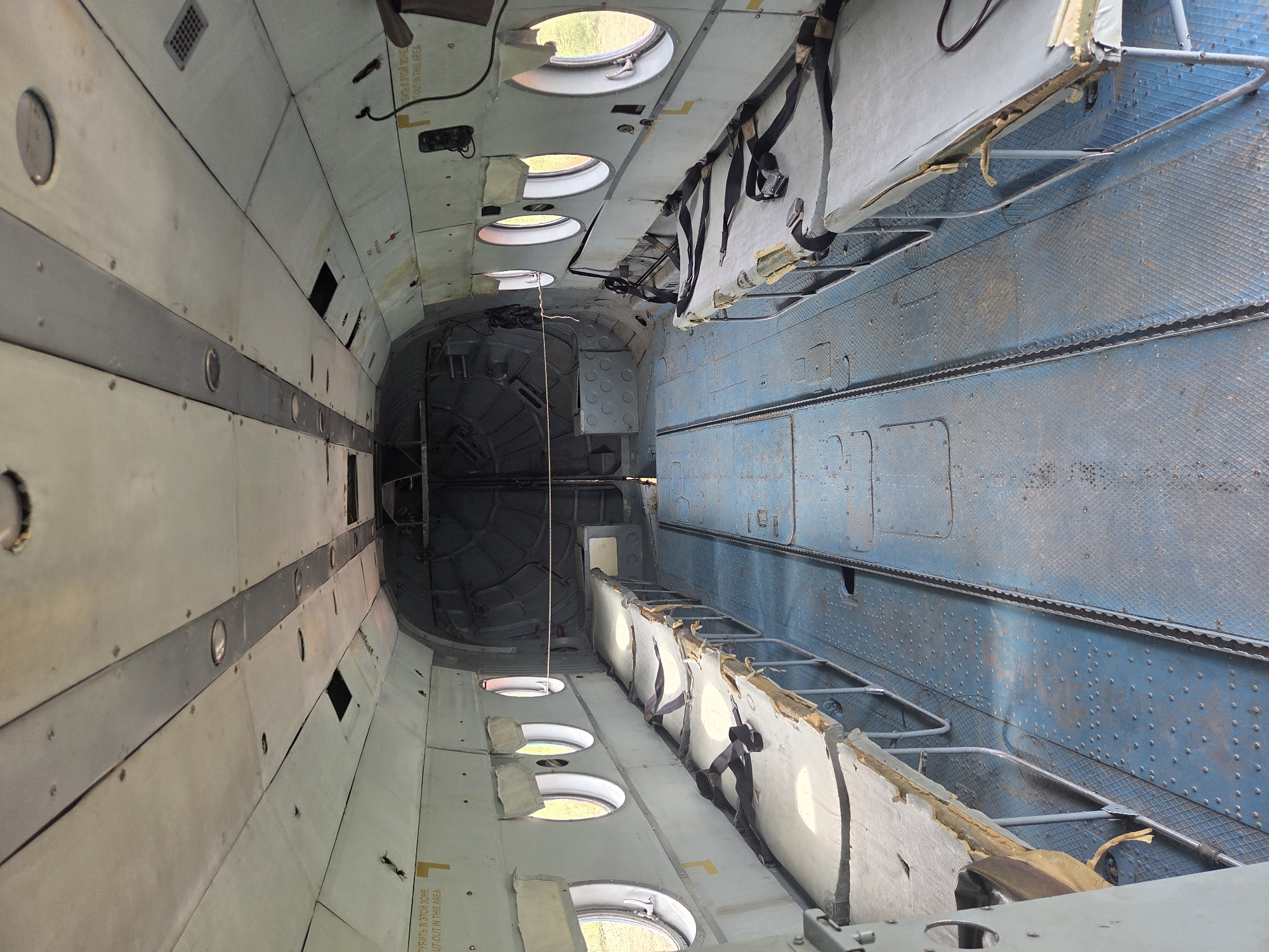 The image shows the interior of a utilitarian aircraft, possibly a former medical evacuation aircraft, with rows of cots on either side of a central aisle. The cots are simple metal frames with worn, padded surfaces and black restraining straps. The floor is a durable blue metal with a patterned surface and a central channel. The walls are a light color, showing signs of age and some damage, with small circular windows spaced along the sides. The ceiling is also light-colored and features recessed lighting fixtures and ventilation grates. The rear of the cabin appears to be a large open cargo area with metal structures. Warning labels with Russian text are visible on the walls, including one that translates to "CUT OUT IN THIS AREA." The environment outside the windows appears to be a grassy, open field. The image does not contain any people or specific indicators of its exact location within Zhuchki, Russia.