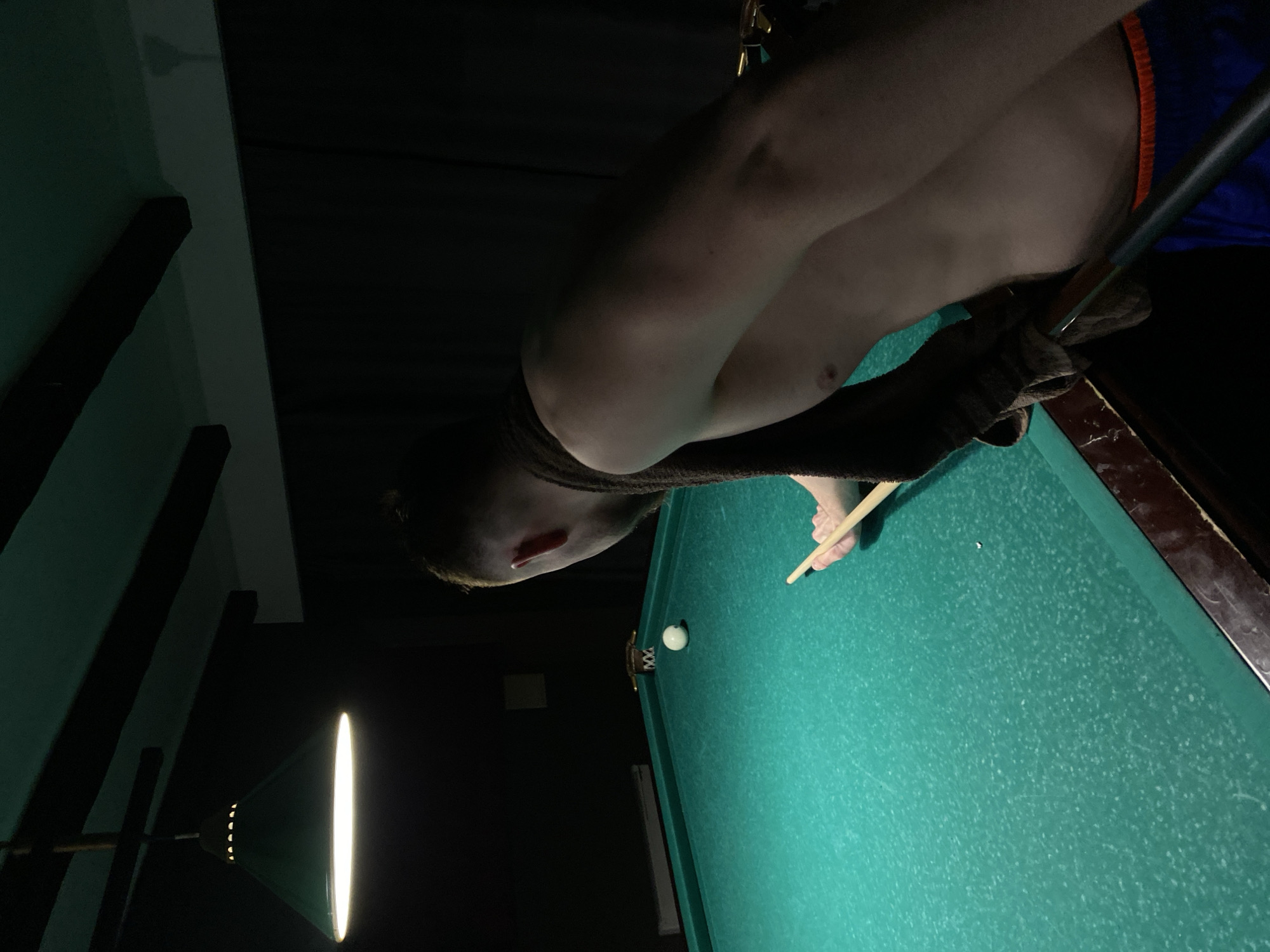 An indoor scene within Perm, Russia, displays a male individual playing billiards under focused illumination. The primary light source is a single, dark green conical pendant lamp suspended from a dark ceiling with visible wooden beams. The surrounding environment is dimly lit, with dark walls, possibly covered by drapes.
The male individual is observed from his upper back and right side, leaning over a green-felted pool table. He is shirtless, with a dark brown towel draped across his right shoulder. Below his torso, blue shorts with an orange waistband are visible. His right hand holds a wooden pool cue, positioned to strike a white cue ball situated on the table. The green felt of the table surface exhibits visible dust and wear. A second, darker billiard ball is partially visible further down the table near a corner pocket. A brass-colored or metallic detail is also present near this distant pocket. The individual's posture indicates concentration on the shot.