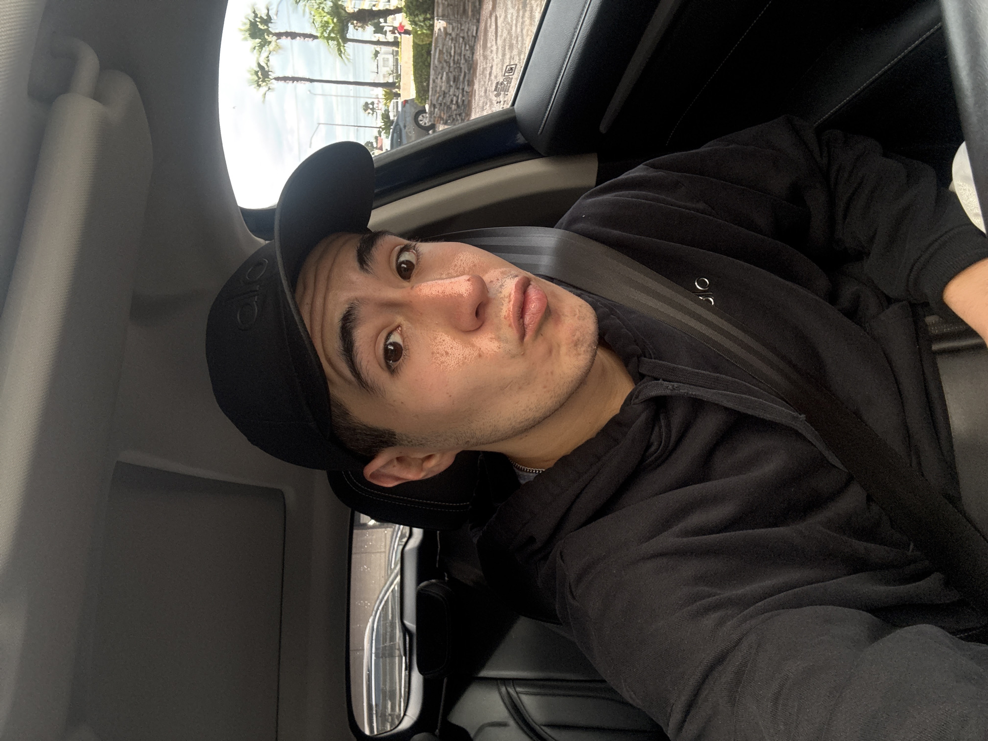 A young man wearing a black baseball cap and a black hoodie is sitting in the driver's seat of a car. He is looking at the camera with his lips pursed. The car appears to be parked, with a view of palm trees and a cloudy sky through the windows. The interior of the car is visible, including the dashboard, sunroof, and black leather seats. The man has a chain necklace visible. The location is specified as San Bernardino Tlaxcalancingo, Mexico.