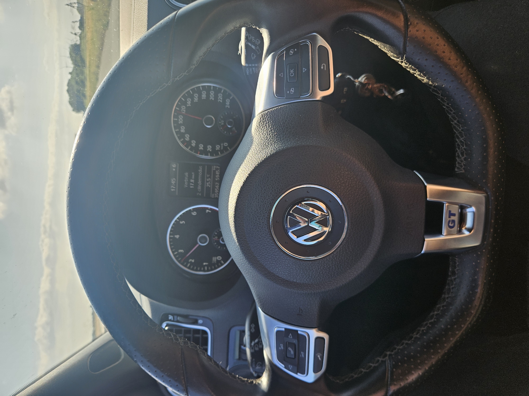 The image is a close-up, interior shot of a black leather steering wheel in a Volkswagen car. The steering wheel has silver accents and controls for the phone and audio system on the left side, and controls for the infotainment system and windshield wipers on the right. The Volkswagen logo is prominently displayed in the center of the steering wheel. The dashboard features a speedometer, tachometer, and a digital display showing the time, fuel consumption, and mileage. Outside the car, a rural landscape with windmills and trees is visible through the windshield under a bright, cloudy sky. The image appears to have been taken while the car was in motion. The location is identified as Meteren, Netherlands.