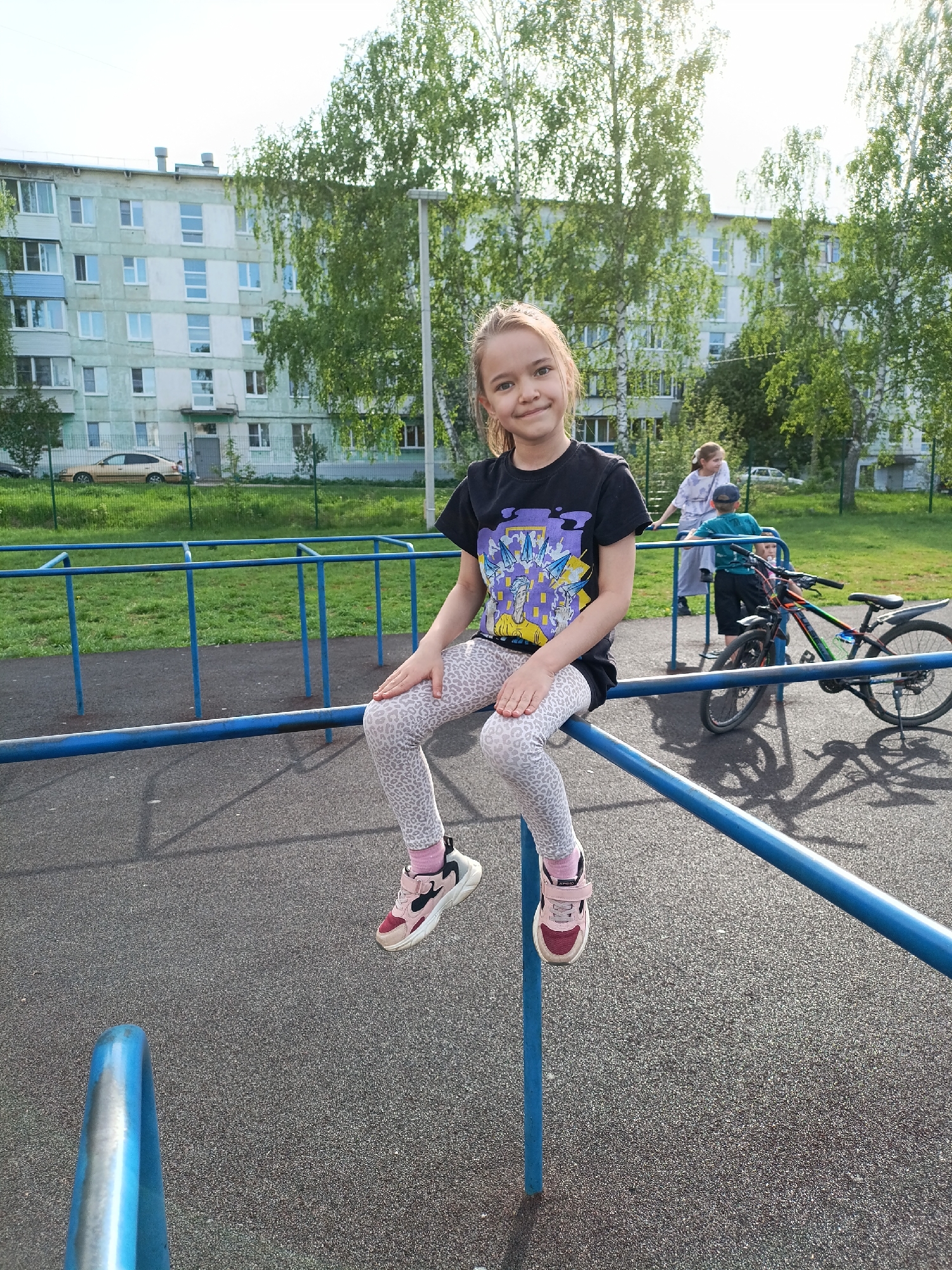 A girl sits on a blue metal rail in a playground, with a residential building, trees, and other people in the background.
Specifically, the girl, likely a child, is sitting on the top rail of what appears to be a climbing structure in a playground. She is wearing a black t-shirt with a graphic design, patterned grey leggings, and pink sneakers. She is facing the camera and smiling. The ground beneath her is asphalt.
In the background, there is a multi-story residential building with many windows. Several tall trees are present. To the right, behind the climbing structure, two adults and a child are standing beside a bicycle. A low metal fence separates the playground from a grassy area. A car is also visible in the background.
Based on visual clues such as the architecture and environment, the location is likely Chaikovsky, Russia.