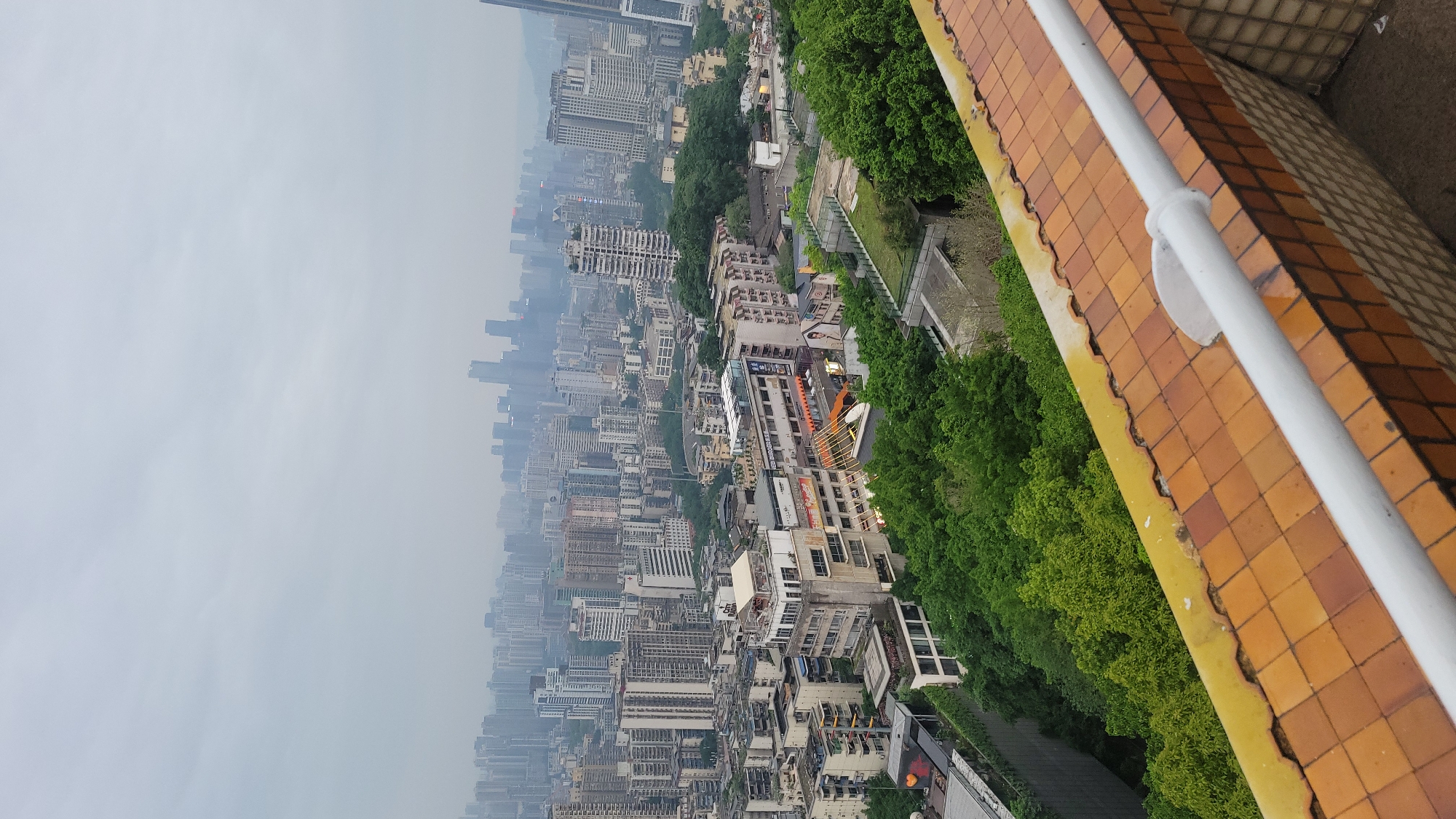 The image presents an elevated view of an urban landscape under overcast skies. The city consists of densely packed buildings of varying heights, merging into a hazy distance. A green belt of trees bisects the city's construction. The foreground features a tiled surface, with a white railing.