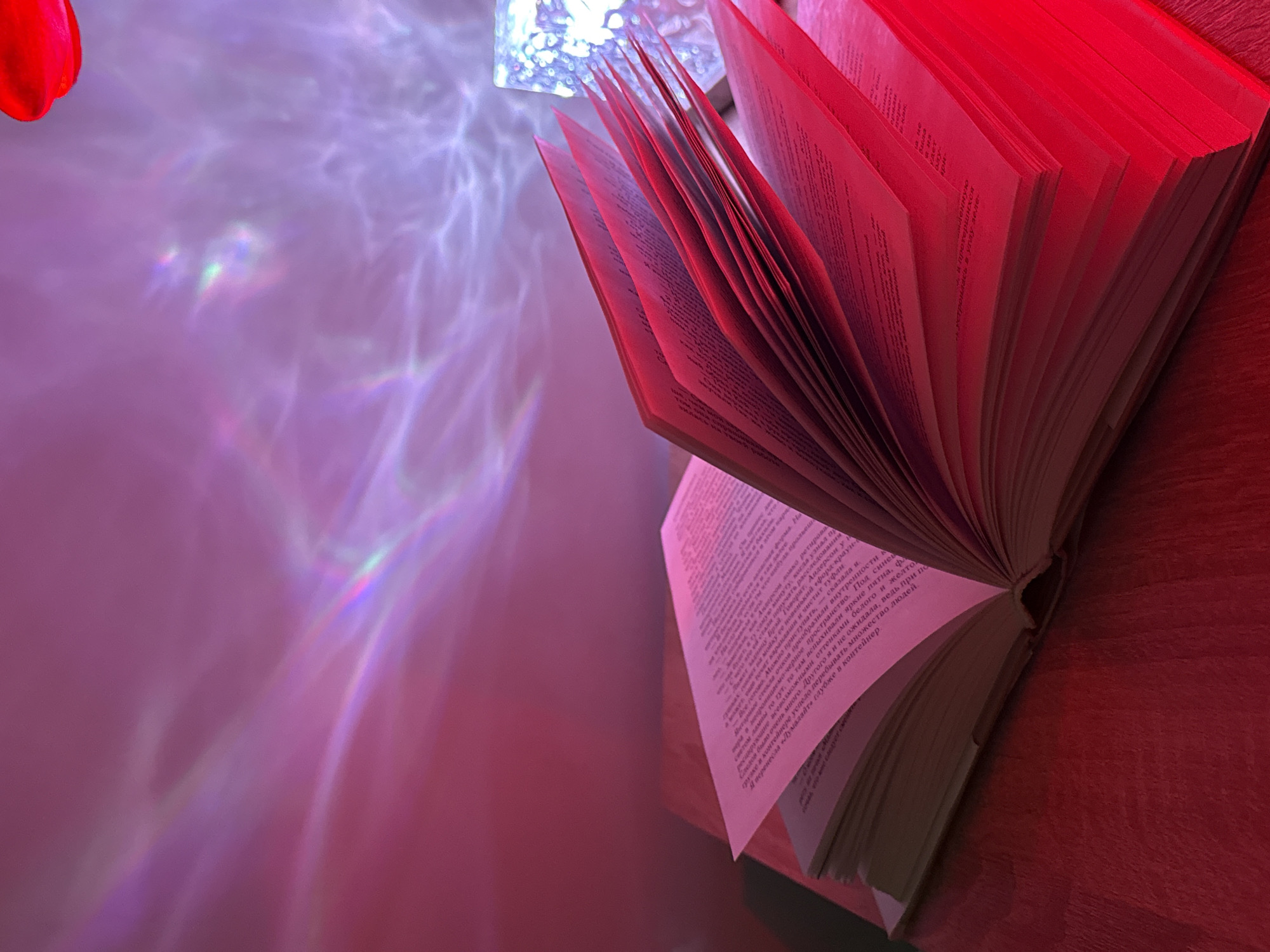 This indoor image features an open book bathed in vivid red and purple light, creating a dramatic and artistic atmosphere. The primary subject is a thick, hardcover book resting on a reddish-brown wooden surface, likely a table or shelf. Its pages are fanned wide open, revealing text that glows red due to the intense lighting.
To the left of the book, a textured light source, possibly a decorative lamp or crystal, projects intricate, wavy patterns of purple, pink, and white light onto a light-colored wall. These light patterns exhibit subtle iridescent or rainbow-like refractions, adding to the ethereal quality of the scene. In the upper left corner of the wider view and one of the cropped images, a small, bright red object, possibly a flower petal or a part of another decoration, is partially visible.
The scene suggests a cozy, contemplative setting, likely in the evening or at night, given the reliance on artificial and colored lighting. There's an implied activity of reading or perhaps simply enjoying the ambiance.
The visible text from the book is primarily in Cyrillic, likely Russian. Fragments suggest a narrative, including phrases such as "will pour and so on," "natural oil form," "if you pour someone," "glasses transformed the insides," "faith in indelible black space," "there flashed bright spots," "Anderson says," "all new 'Ford Crown'," "I moved 'Lumalayte deeper into the container'," "managed to visit many people," and "There were a lot of traces." These snippets hint at a story, possibly a modern narrative or even a science fiction context with names like "Anderson" and products like "Lumalayte" and "Ford Crown."