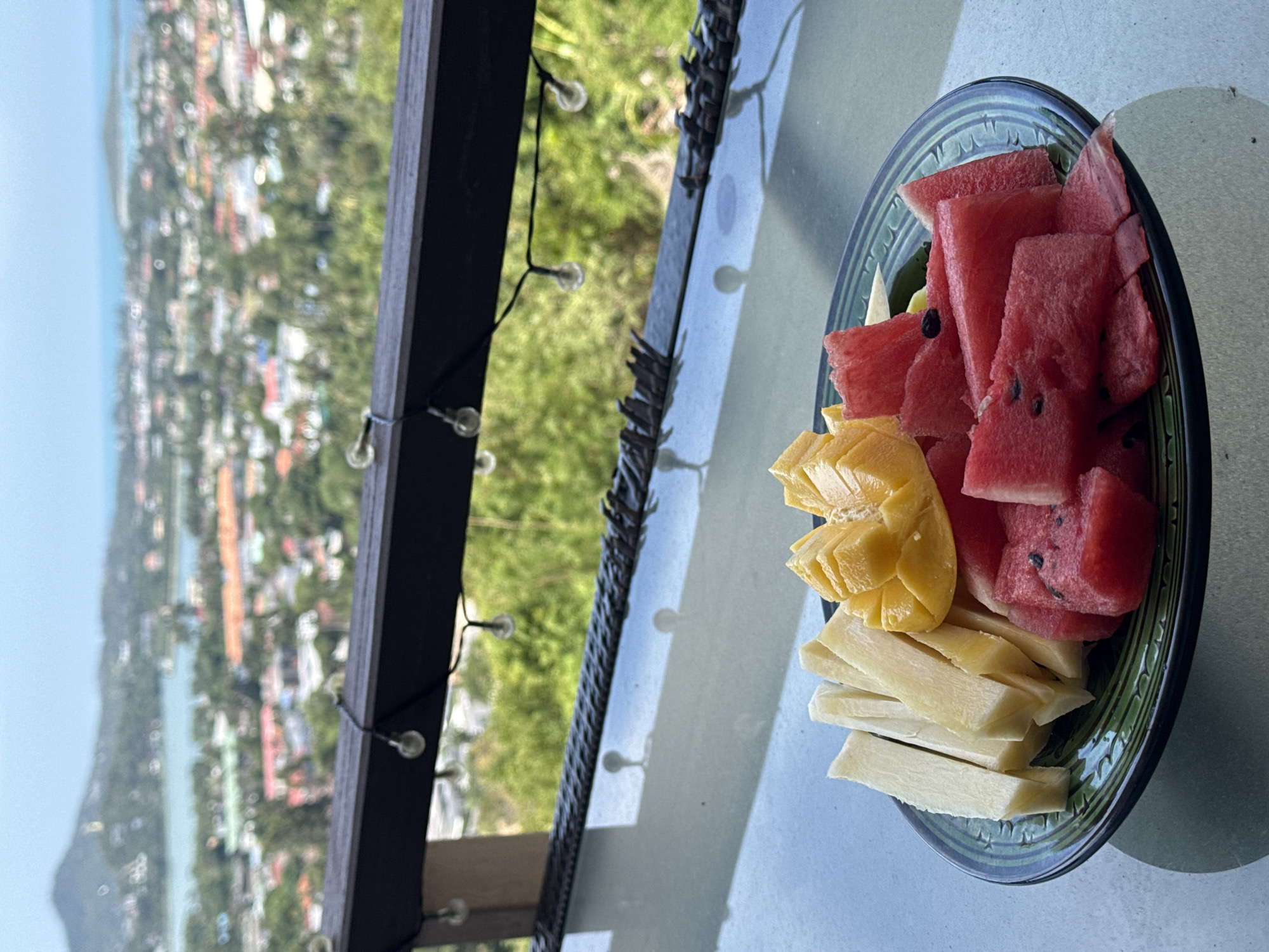 A platter of chopped fruit, including watermelon and mango, rests on a glass-topped table. The platter is dark green with an etched pattern. In the background, out of focus, is a view of a city with buildings and trees, leading to a body of water and a distant hillside under a hazy sky. String lights are visible along a dark railing above the table. This scene appears to be from a balcony or an elevated vantage point in Ko Samui District, Thailand.