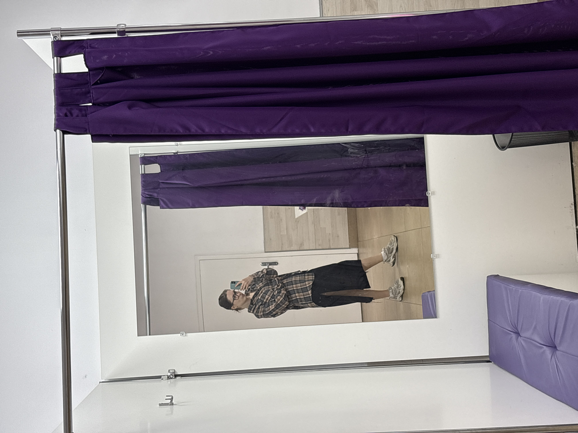 A young woman with dark hair pulled back and wearing glasses is standing in a fitting room, taking a mirror selfie. She is dressed in a plaid button-down shirt and a black midi skirt with a slit up the side. She is also wearing athletic shoes. The fitting room has white walls, a mirrored wall, and a purple curtain. There is a purple bench in the foreground and a trash can visible near the curtain. The floor is tiled. The image appears to have been taken in Saint Petersburg, Russia, given the overall aesthetic of the fitting room and the style of the clothing.