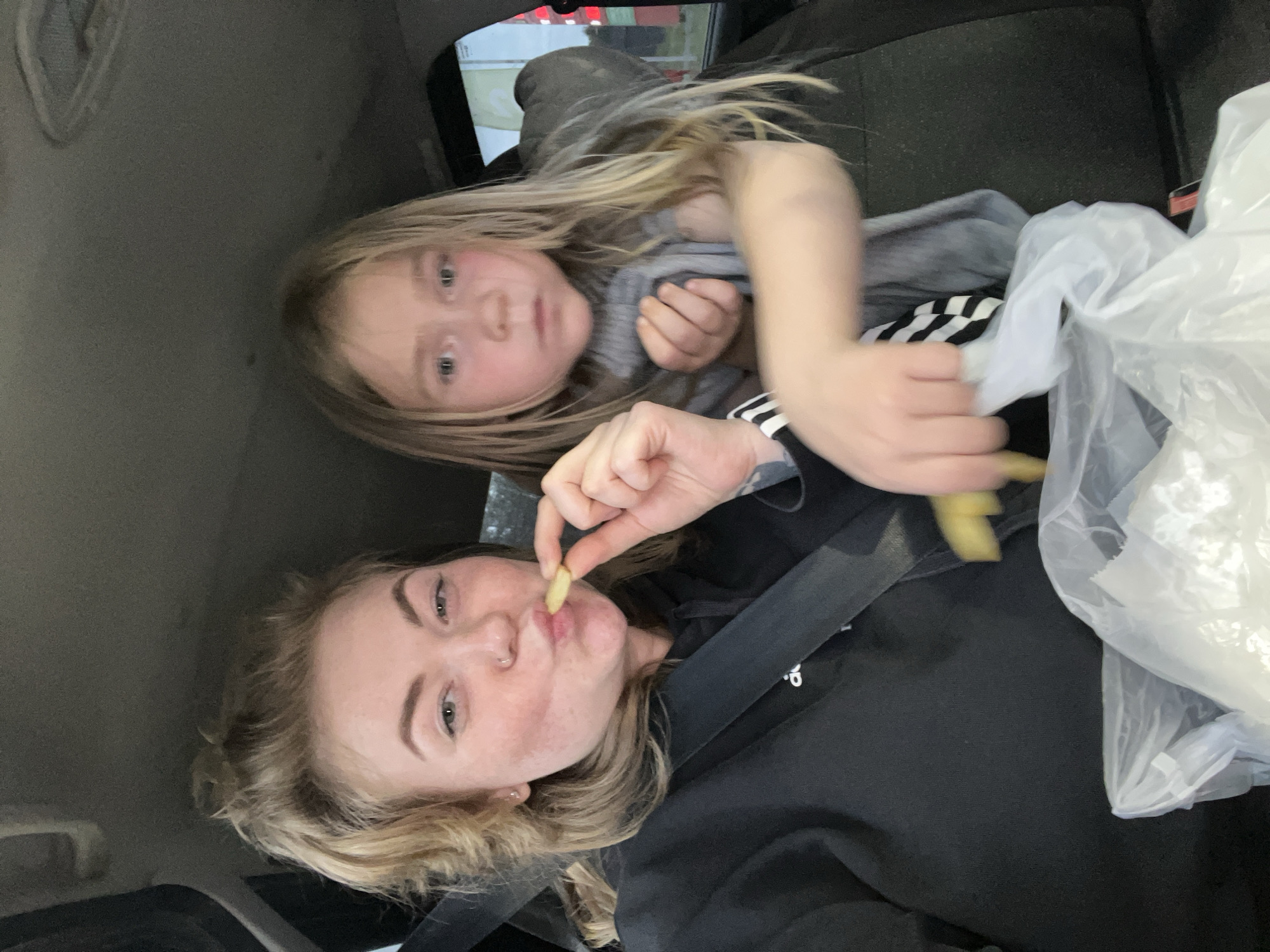 This media file shows two people in what appears to be the interior of a car. The subject in the foreground is a young woman with blonde hair and freckles, wearing a black hoodie and a seatbelt. She is holding a french fry up to her mouth, with a playful expression. To her left and slightly behind her is a young girl, also with blonde hair. She is wearing a grey sweater and appears to be looking at the camera. Next to the woman, there is a white plastic bag. The scene suggests a casual outing, possibly a drive-thru experience or a snack break during a car ride. The lighting appears to be natural, indicating it's daytime. There are no visible text elements in the image. The overall mood is lighthearted and candid.