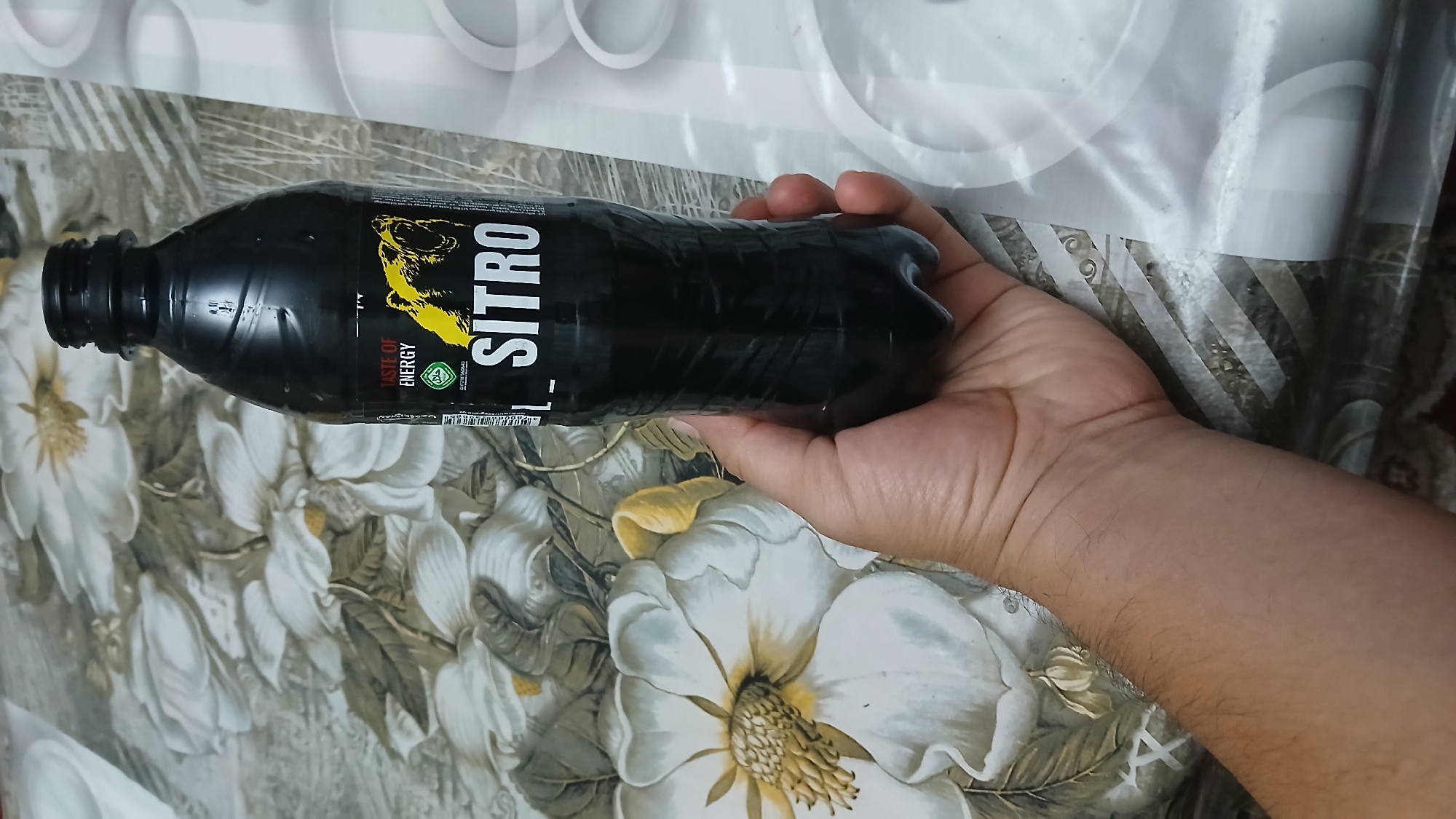 A hand holds a black plastic bottle of "SITRO" energy drink. The bottle has a yellow bear logo and the text "TASTE OF ENERGY" on its label. The background is a patterned tablecloth with floral motifs. The scene is indoors, on a table. There is no identifiable urban or geographical context visible to suggest a specific location in Kokand City, Uzbekistan.
