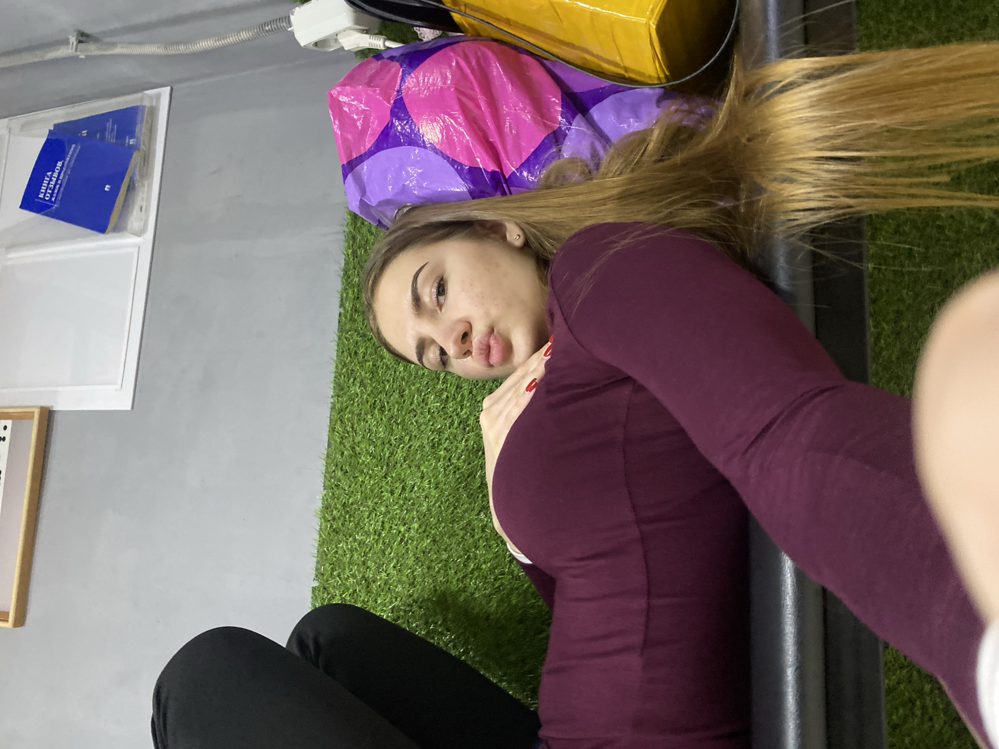 A young woman with long, blonde hair lies on her back on a bench covered with artificial turf. She is wearing a dark purple, long-sleeved shirt and black pants. Her right hand is resting on her chest, with red nail polish visible. She is looking at the camera with her lips pursed. To her right are two large, crinkled plastic bags, one purple and pink, and one yellow. In the background, a grey wall is visible, with a framed picture and a blue book in a clear holder mounted on it. The overall setting appears to be indoors, possibly a waiting area or a break room.