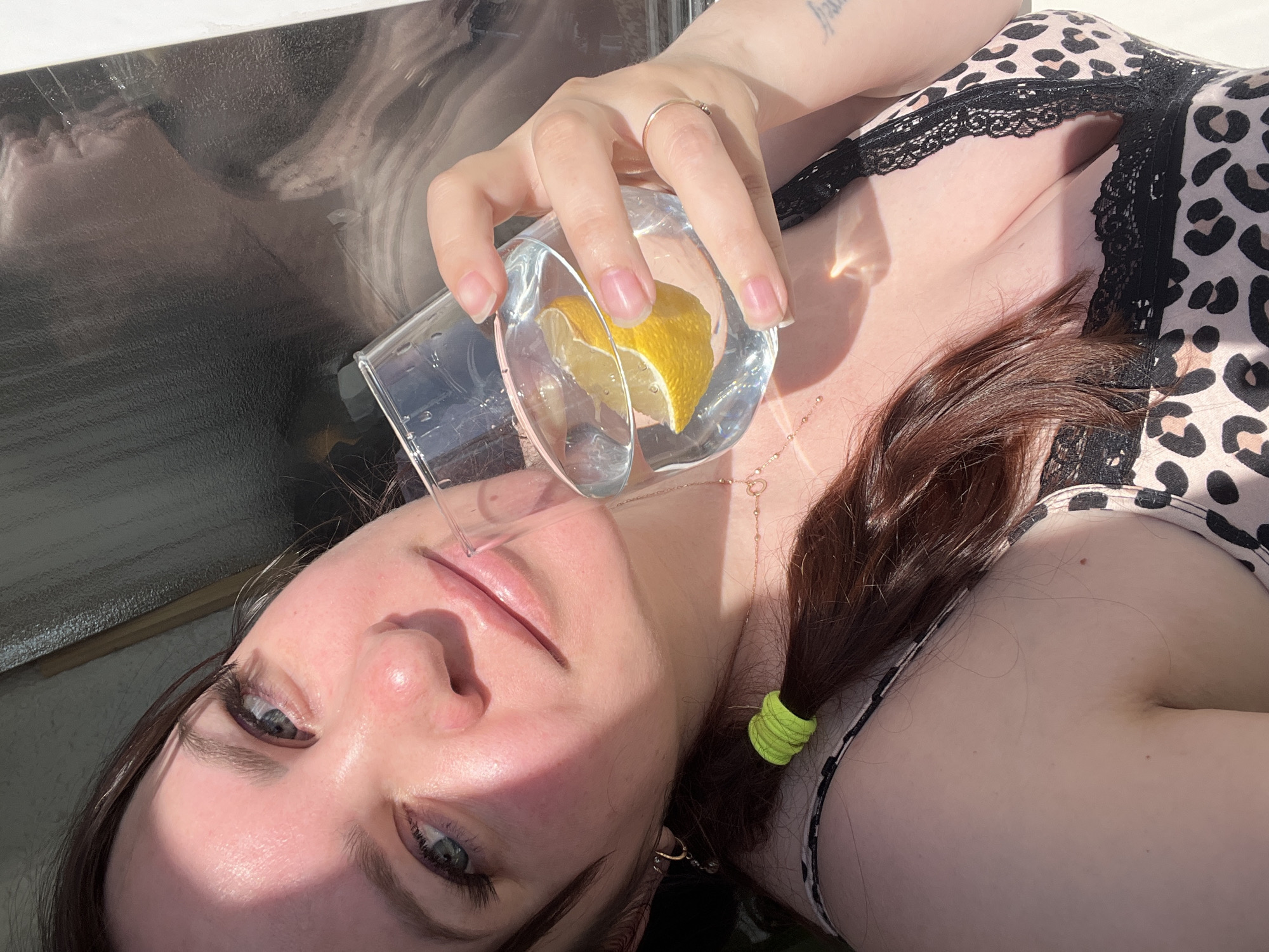 A woman lies in the sun, holding a glass of water with a lemon slice in it. She has brown hair pulled back with a green elastic, fair skin, and is wearing a leopard print top with black lace trim. She wears a ring on her left hand and a necklace. The woman's eyes are blue-gray. Part of what appears to be a reflective surface is to the left of her face. The location in the city of Noviy, Russia is unknown.