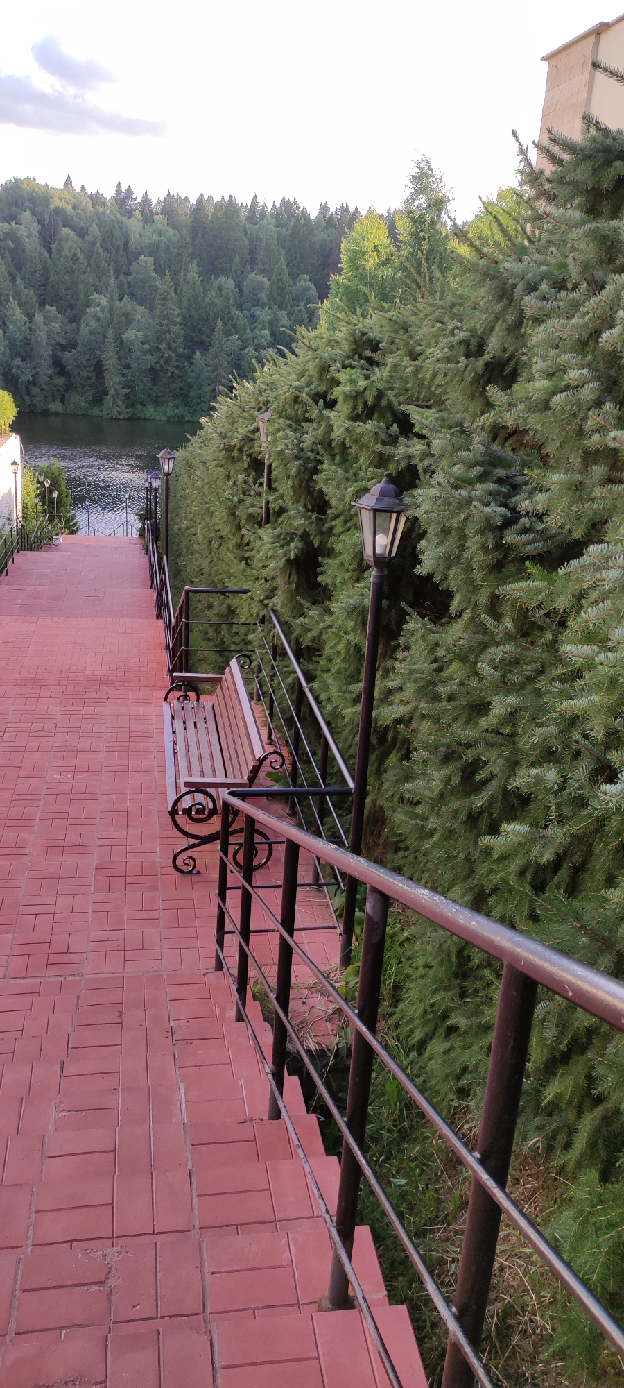 An outdoor scene depicts a red brick pathway descending a staircase, flanked by a dark metal railing and dense evergreen trees. A wooden bench with decorative metal legs is situated on the landing of the stairs. Several old-fashioned street lamps are visible along the path. In the background, a calm body of water is bordered by a lush, green forest under a bright, overcast sky. The overall setting suggests a park or recreational area within a suburban or rural landscape.