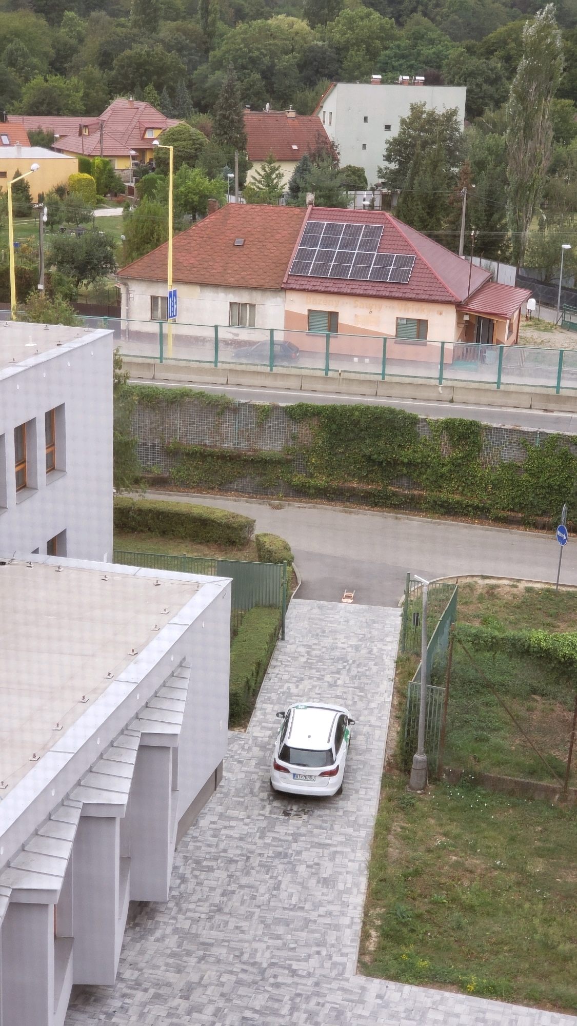 The image depicts an aerial view of a residential area in Prešov, Slovakia, with a focus on modern architecture and infrastructure. A white car is parked on a paved driveway in front of a modern building with a light-colored facade. The building features a sloped, silver-colored roof with visible metal fixtures. Adjacent to this structure, a neighboring house with a red-tiled roof is partially visible, distinguished by solar panels installed on its rooftop. The surrounding environment includes a paved road, a fence line, and dense green foliage, characteristic of a suburban or semi-rural setting. The weather appears overcast.
