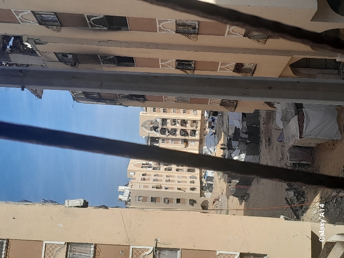 The image captures a damaged urban environment in Khan Yunis, Palestinian Territory, viewed from an elevated position. Multiple multi-story residential buildings, primarily light beige in color with brown accents, feature decorative arched window frames and geometric patterns. The building on the immediate right exhibits substantial damage to its upper sections, with exposed rebar and fractured concrete visible; many of its windows are open or broken, some retaining metal grilles. Similar damage is evident on other visible structures.
In the foreground, several dark, elongated vertical objects and a lighter concrete pillar partially obstruct the view. Between the buildings, a large, unpaved expanse of dirt and rubble is visible. This area is densely populated with numerous makeshift shelters and tents constructed from tarpaulins, fabrics, and various salvaged materials. Laundry is observed hanging outside some of these temporary structures. A small group of approximately three to four individuals is discernible in the mid-ground, walking on the unpaved surface amidst the shelters. The sky above is clear blue. A "Galaxy A14" watermark is present in the bottom-left corner.