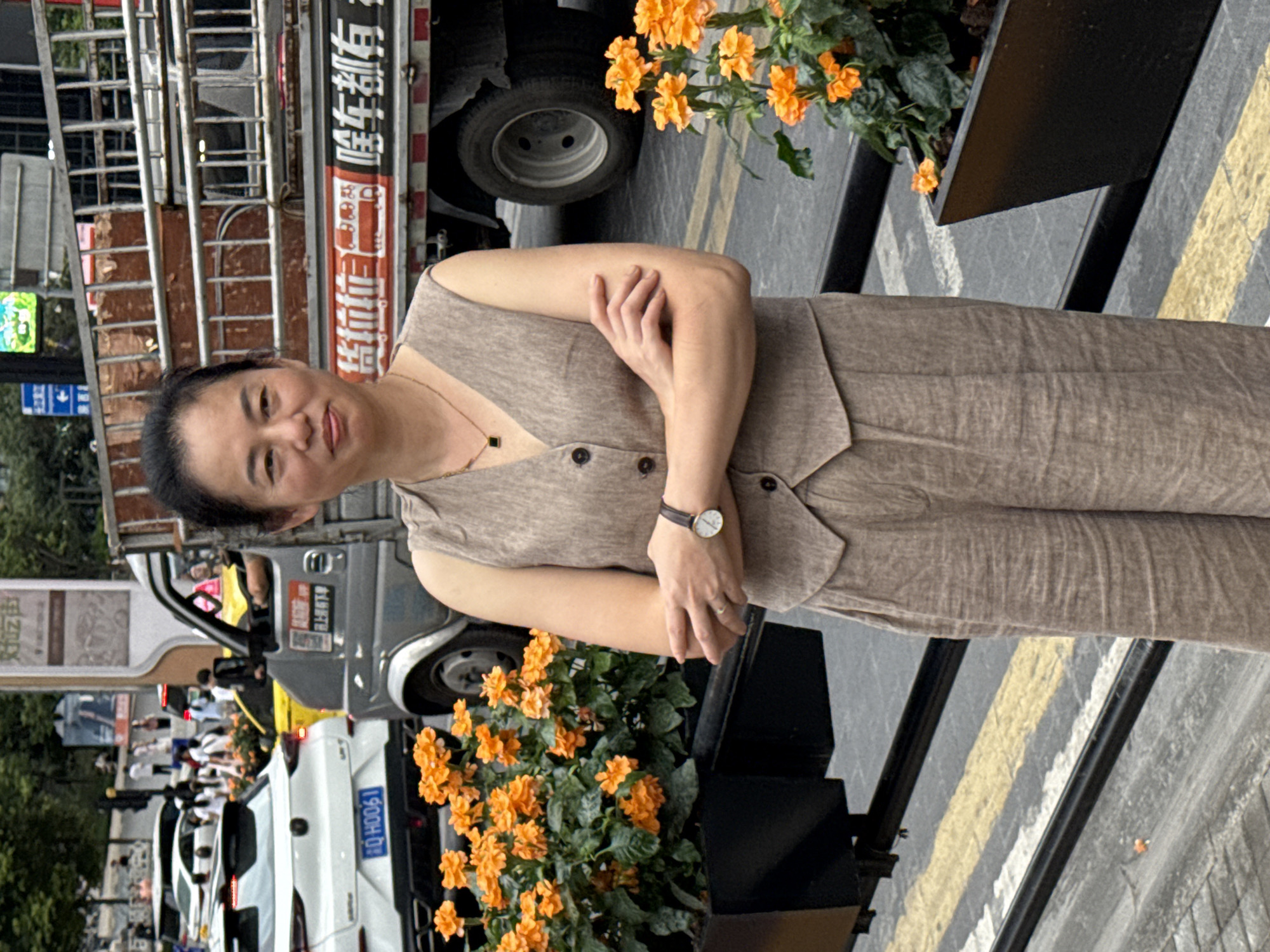 A full-shot, outdoor, eye-level, vertical, medium-light exposure photograph captures a middle-aged East Asian woman standing in front of a parked truck and some orange flowers in Yuzhong District, China. The woman, with dark hair pulled back and fair skin, is wearing a light brown sleeveless vest with two buttons and matching wide-leg pants. She has her arms crossed and is looking towards the camera with a slight smile. A gold necklace with a black square pendant is visible, as is a wristwatch on her left wrist. The background features a truck with a metal ladder rack, its side displaying Chinese characters, and a white car with a license plate reading "RQ HO061". Orange flowers in dark rectangular planters are on the left and right sides of the frame. The ground is paved, with yellow and white lines indicating a crosswalk. The overall scene suggests a street or roadside setting in an urban environment.