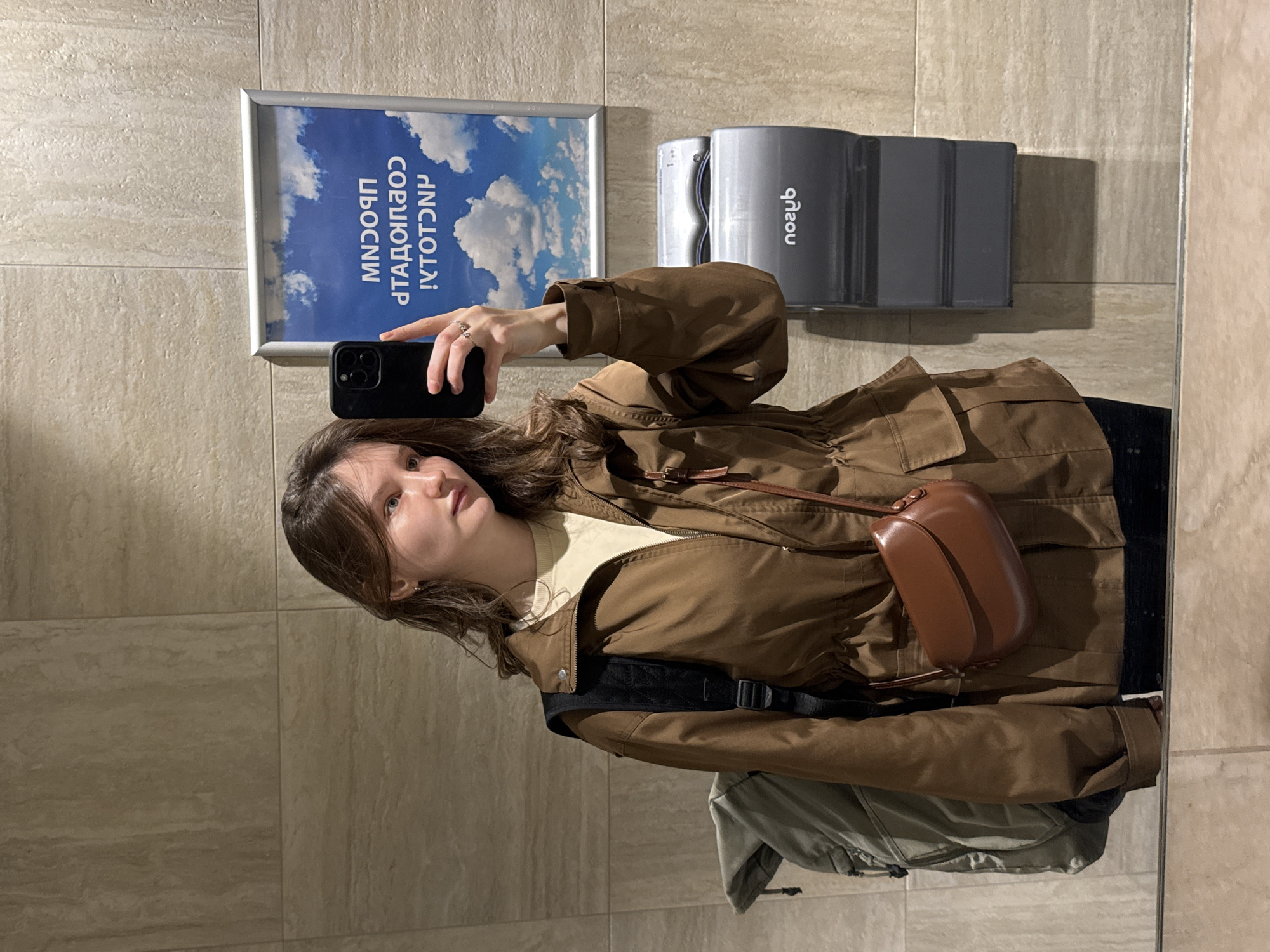 A woman is taking a selfie in what appears to be a public restroom. She is holding a black smartphone in her right hand and looking at the screen. She is wearing a brown jacket, a cream-colored top, and a small brown purse is slung across her body. A black backpack strap is visible on her shoulder, and what looks like a green jacket or coat is partially visible below her jacket. The background consists of a tiled wall, a framed picture of a blue sky with clouds, and a gray hand dryer. Based on the text in the framed picture, the location is likely in the city of Bolshoye Savino, Russia.