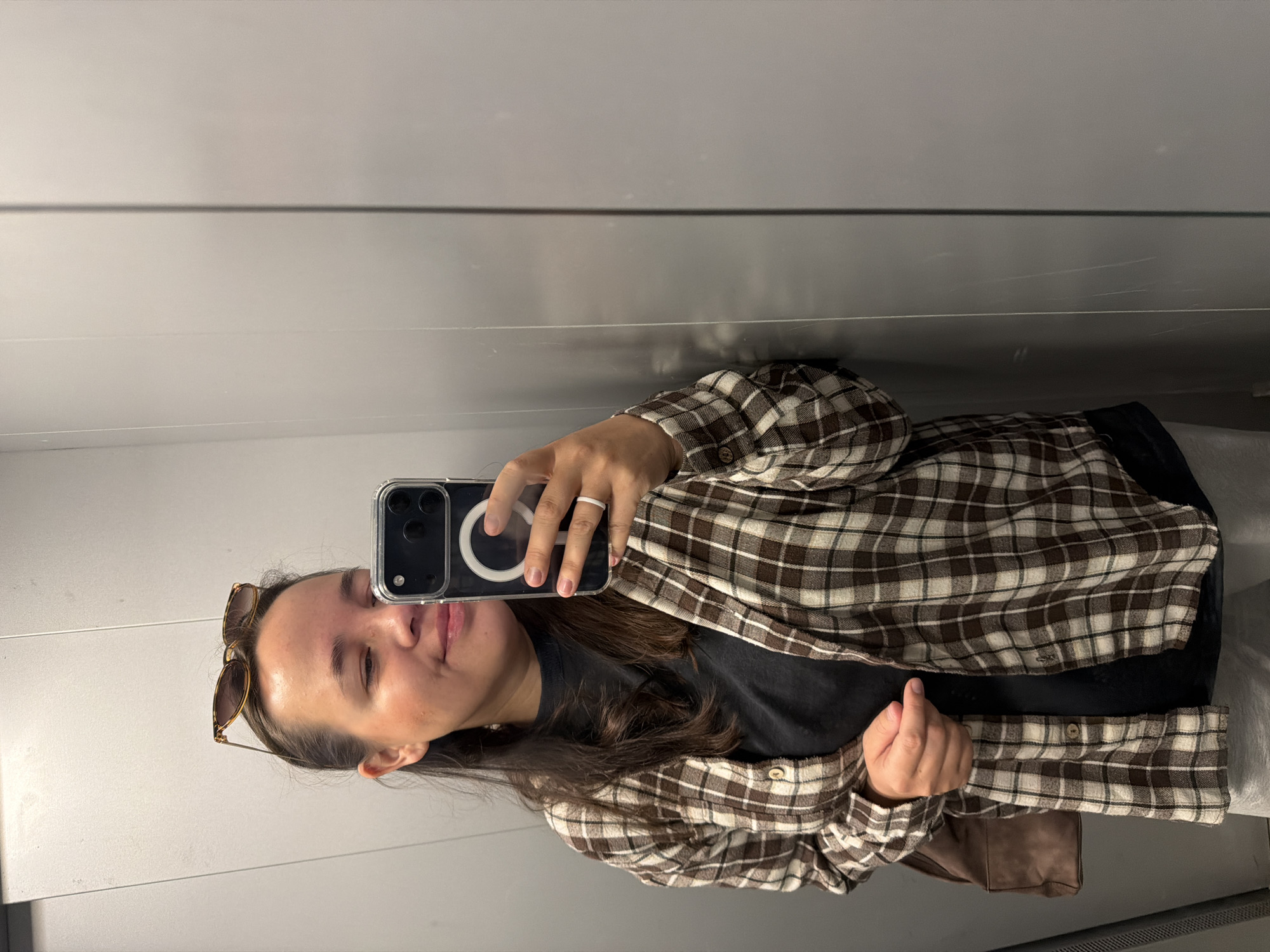 A person takes a selfie in what appears to be a train or bathroom. The person is wearing a brown and white plaid shirt over a dark t-shirt and glasses are perched atop her head. She holds a phone in her right hand. The background appears to be gray paneling. The exact location in Chaikovsky, Russia cannot be determined from the image.