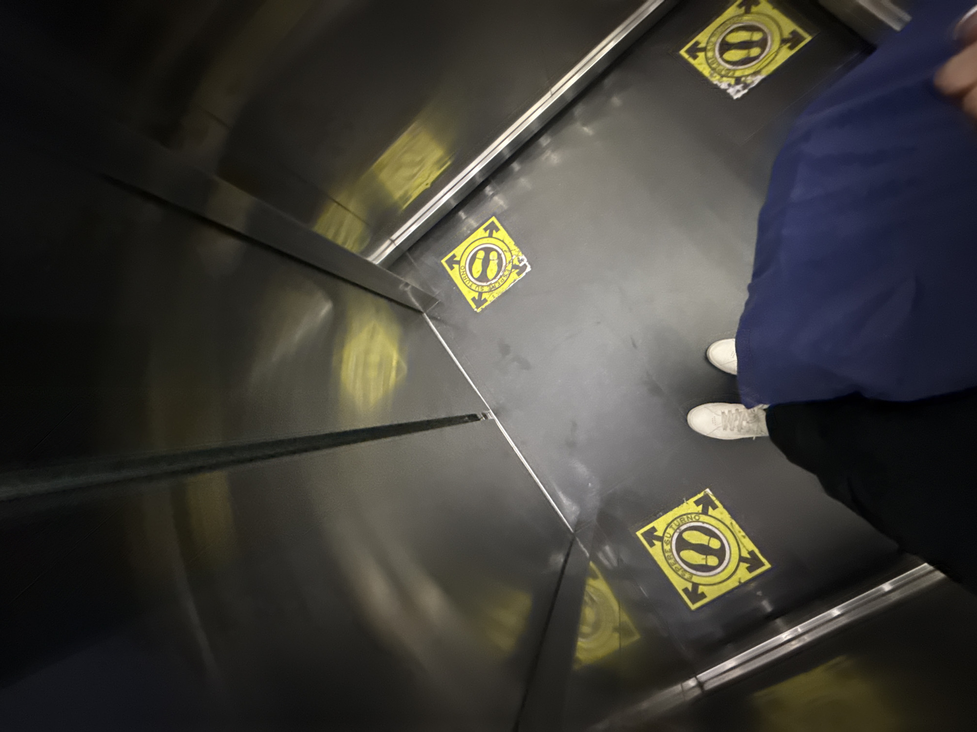 A person stands in an elevator in San Bernardino Tlaxcalancingo, Mexico. The elevator's interior is primarily a polished metal. The floor has multiple square yellow stickers with a graphic of two footprints inside a circle and arrows pointing in four directions, with the text "ESPERE SU TURNO" bordering the square. The person wears white sneakers, dark-colored pants, and a dark blue upper garment.