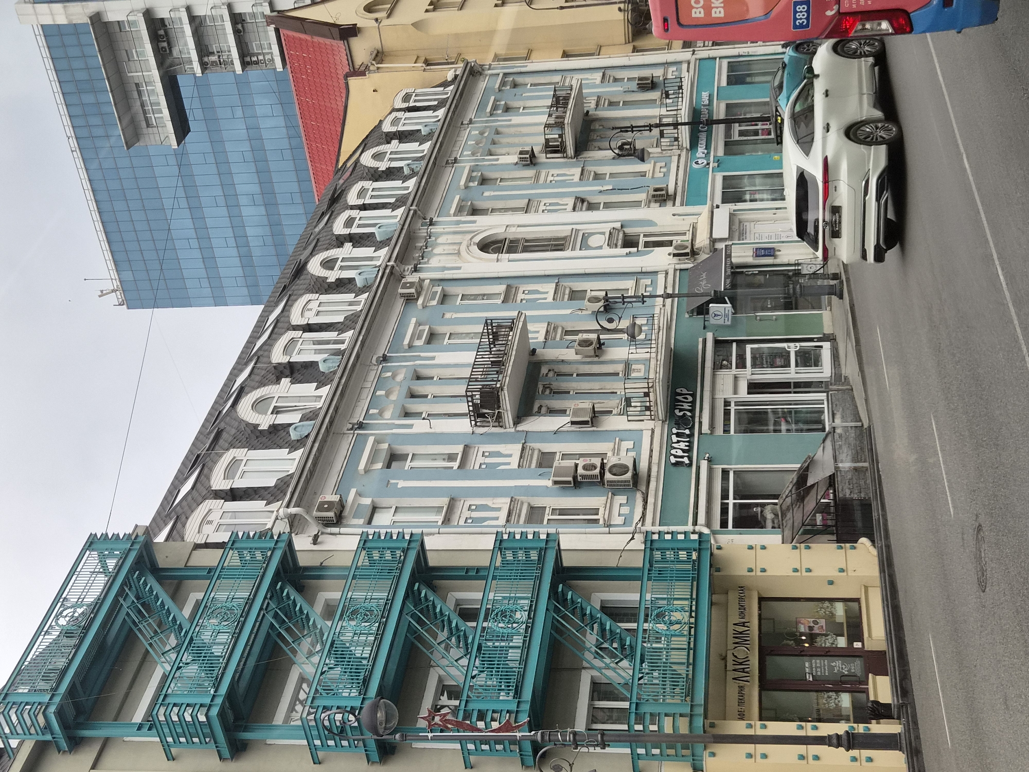 The image shows a street view in Vladivostok, Russia. A multi-story blue and white building with ornate architectural details and a mansard roof is prominent in the foreground. A teal-colored exterior staircase runs up the side of an adjacent building. Several shops with signs in Cyrillic script are visible on the ground floor of the blue and white building. One sign reads "IPATI SHOP". Another shop has text that appears to be a bakery. A white SUV is driving down the street, and the back of a red and blue bus is visible on the right side of the frame. In the background, a modern glass-fronted building rises above the older structures. The sky is overcast.