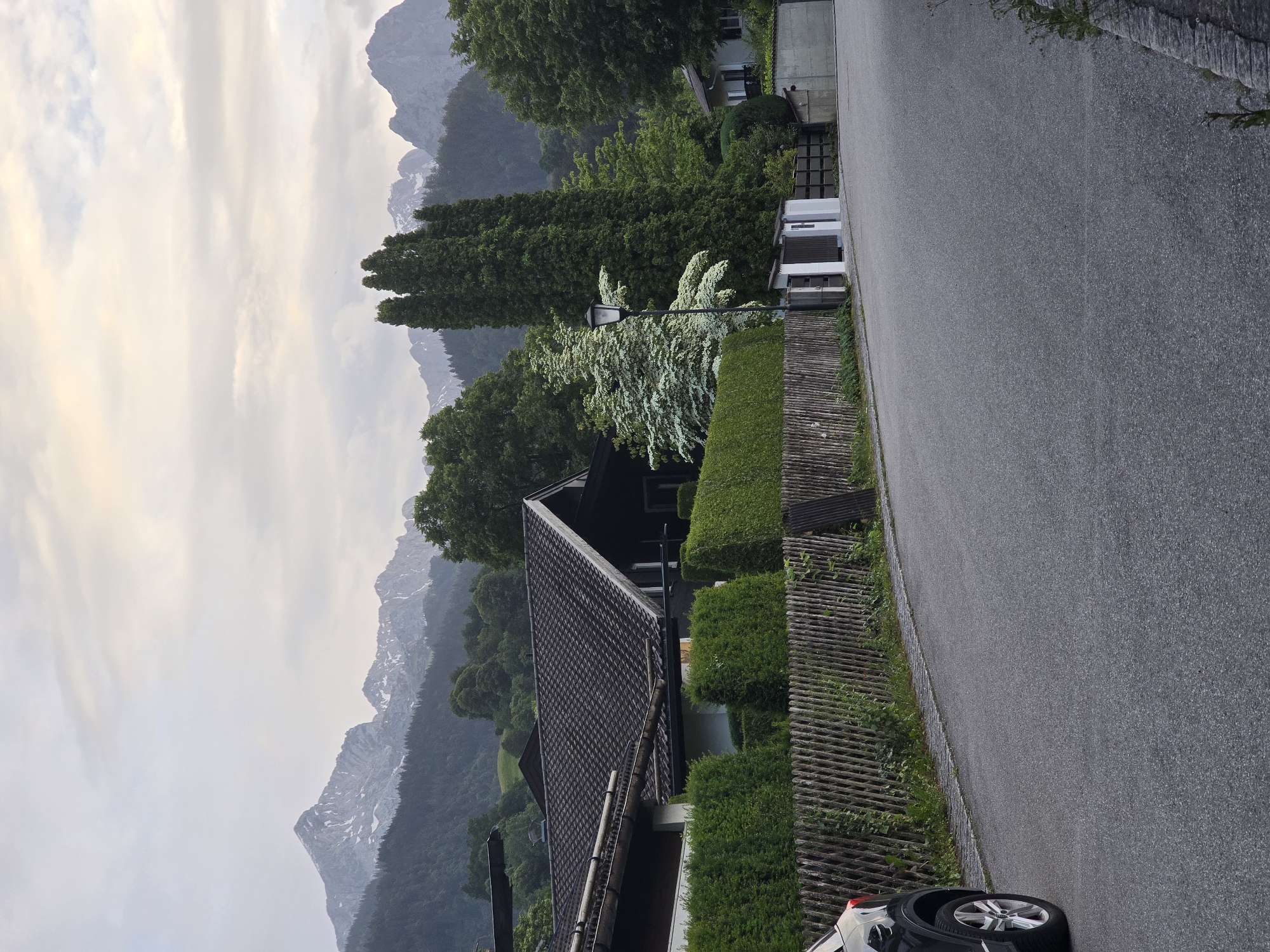 The image depicts a residential street in Garmisch-Partenkirchen, Germany, with a view of snow-capped mountains in the background. The foreground features a paved road with a dark-colored car parked on the left side. A wooden fence runs along the right side of the road, bordering a well-maintained hedge. Beyond the hedge, houses with dark roofs and light-colored walls are visible, set amongst lush green trees. In the mid-ground, tall evergreen trees and a distinctive white-flowering tree stand out. The sky is overcast with a soft, diffused light. The overall scene suggests a quiet, suburban environment in a mountainous region.