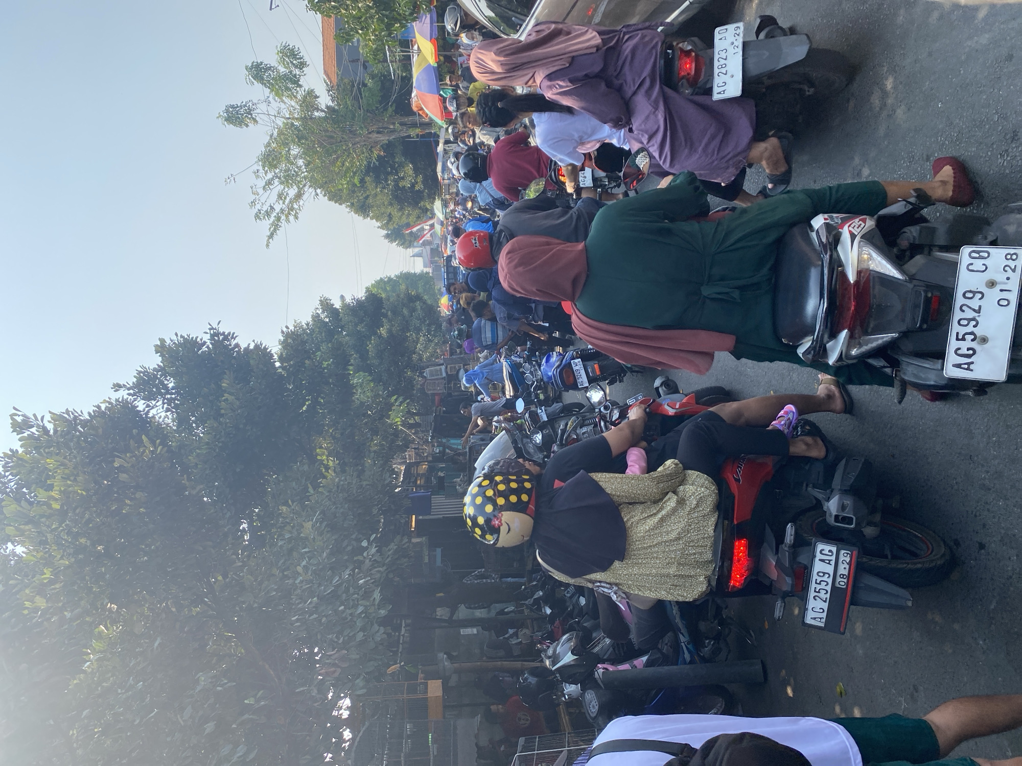 The image captures a bustling street scene in Kediri City, Indonesia, characterized by heavy motorcycle traffic and pedestrian activity, likely at a market or busy thoroughfare. The foreground features several motorcycles with riders, most wearing helmets, with one rider in a distinctive yellow helmet with a dotted pattern. People are dressed in everyday clothing, with many women wearing headscarves in various colors, including red, purple, and green. The background reveals stalls with goods, possibly a market, and more motorcycles and people extending down the street. Trees line the sides of the road, and the sky above is clear and bright, suggesting a sunny day. The overall impression is one of a vibrant and crowded urban environment.