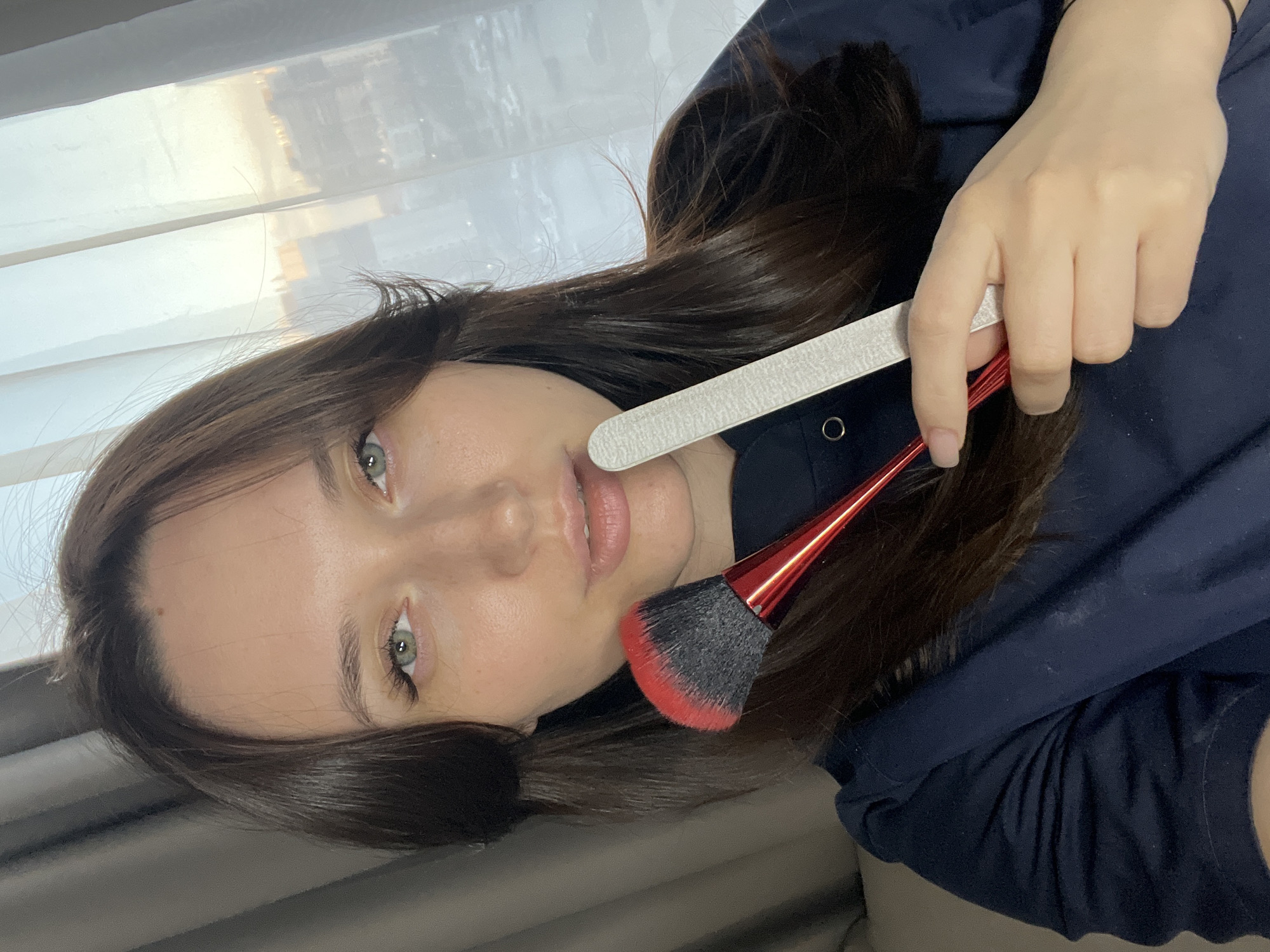 A close-up, rotated portrait features a young woman with long, dark brown hair and striking light green eyes, looking directly at the viewer with a neutral expression. She has fair skin and slightly parted lips. She is wearing a dark blue long-sleeved top. Her right hand is visible in the upper right quadrant, holding a white nail file and a red-handled makeup brush with black and red bristles, suggesting a beauty-related activity. The background consists of a brightly lit window with sheer white curtains or blinds, through which blurred reflections of what appear to be outdoor structures or sky are visible. The natural light from the window indicates it is daytime. There is no visible text in the image. The scene is set indoors, potentially in Tyumen, Russia.