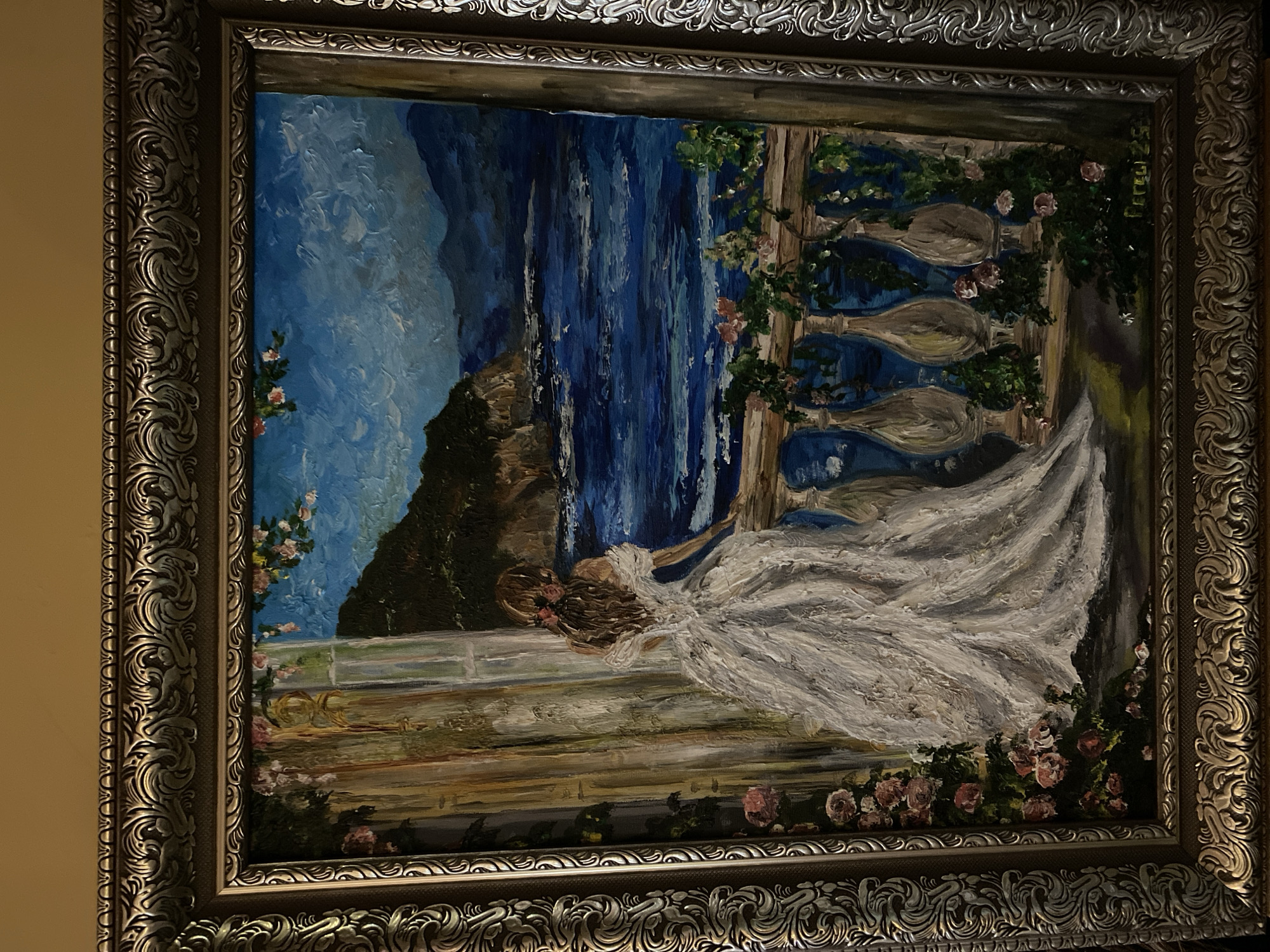 This is an oil painting framed in an ornate silver and gold frame. The painting depicts a woman in a voluminous white gown with her back to the viewer, standing on a balcony overlooking a body of water and mountainous terrain. The woman has long brown hair with a pink ribbon. The balcony features a balustrade with vase-like supports and is adorned with pink roses and green foliage. The seascape is rendered with textured brushstrokes, showing waves and distant mountains under a bright blue sky. The scene is evocative of a romantic, perhaps Mediterranean, setting. There is a signature in the lower right corner that appears to read "Perugi 25". The painting is displayed against a plain, warm-toned wall, suggesting it is likely hanging in a domestic or gallery setting. The location in Poltava, Ukraine is not discernible from the visual information provided.