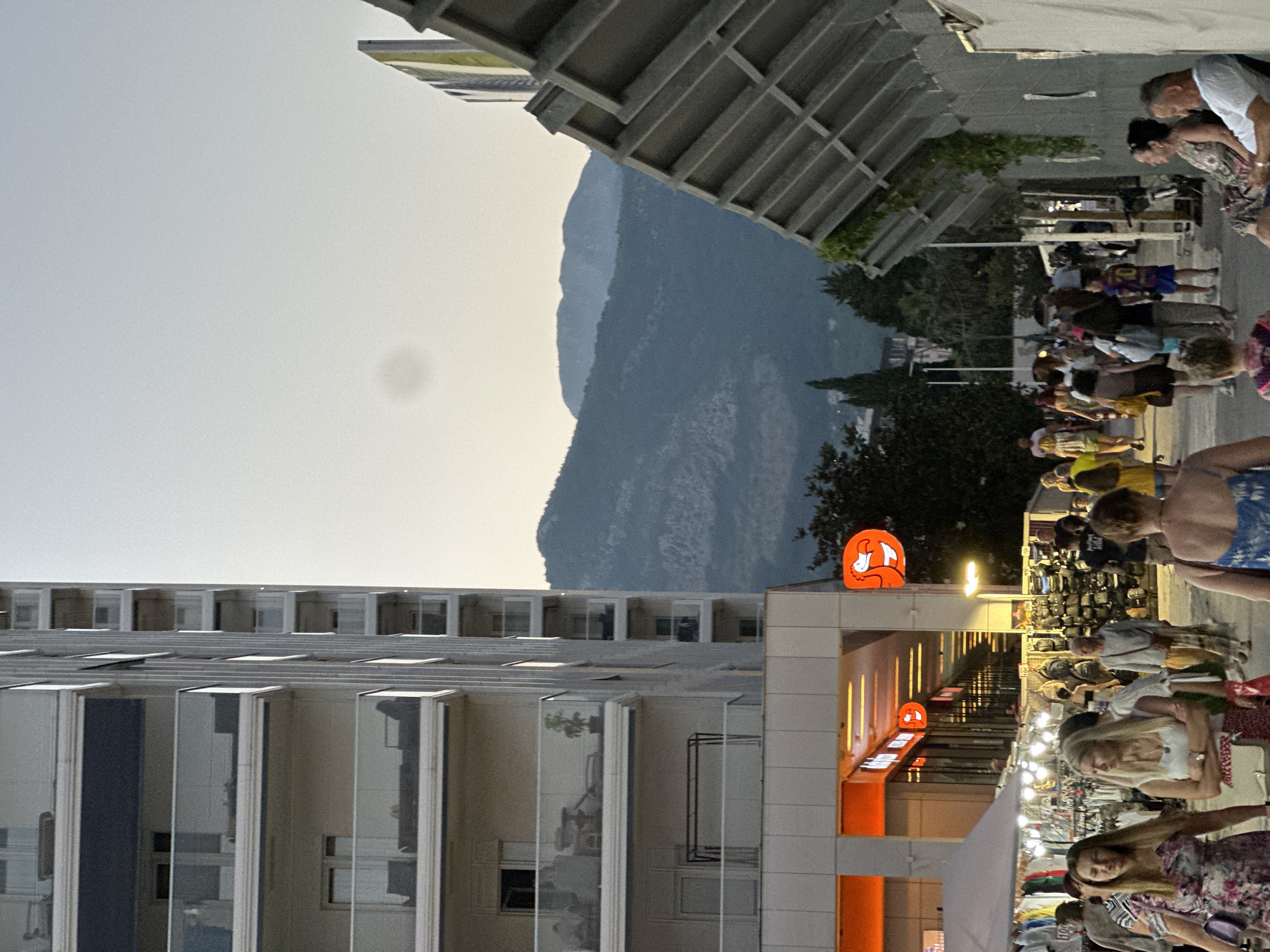 The image depicts a street scene in Budva, Montenegro, likely in the late afternoon or early evening, as indicated by the soft light and shadows. A modern, multi-story apartment building with balconies dominates the left side of the frame. On the right, a textured, angular structure, possibly part of a building or a covered walkway, extends into the foreground.
A bustling pedestrian walkway is visible in the center, populated by numerous people walking in various directions. Some are casually strolling, while others appear to be browsing at market stalls or shops lining the street. Market tents with clothing and accessories are set up on the left side of the walkway. Shopfronts with bright orange illuminated signs are visible behind the tents. A distinctive orange flamingo sign is prominently displayed on the right.
In the background, lush green hills and mountains rise against a pale, hazy sky. The overall impression is one of a lively tourist area with a blend of contemporary architecture and natural scenery.