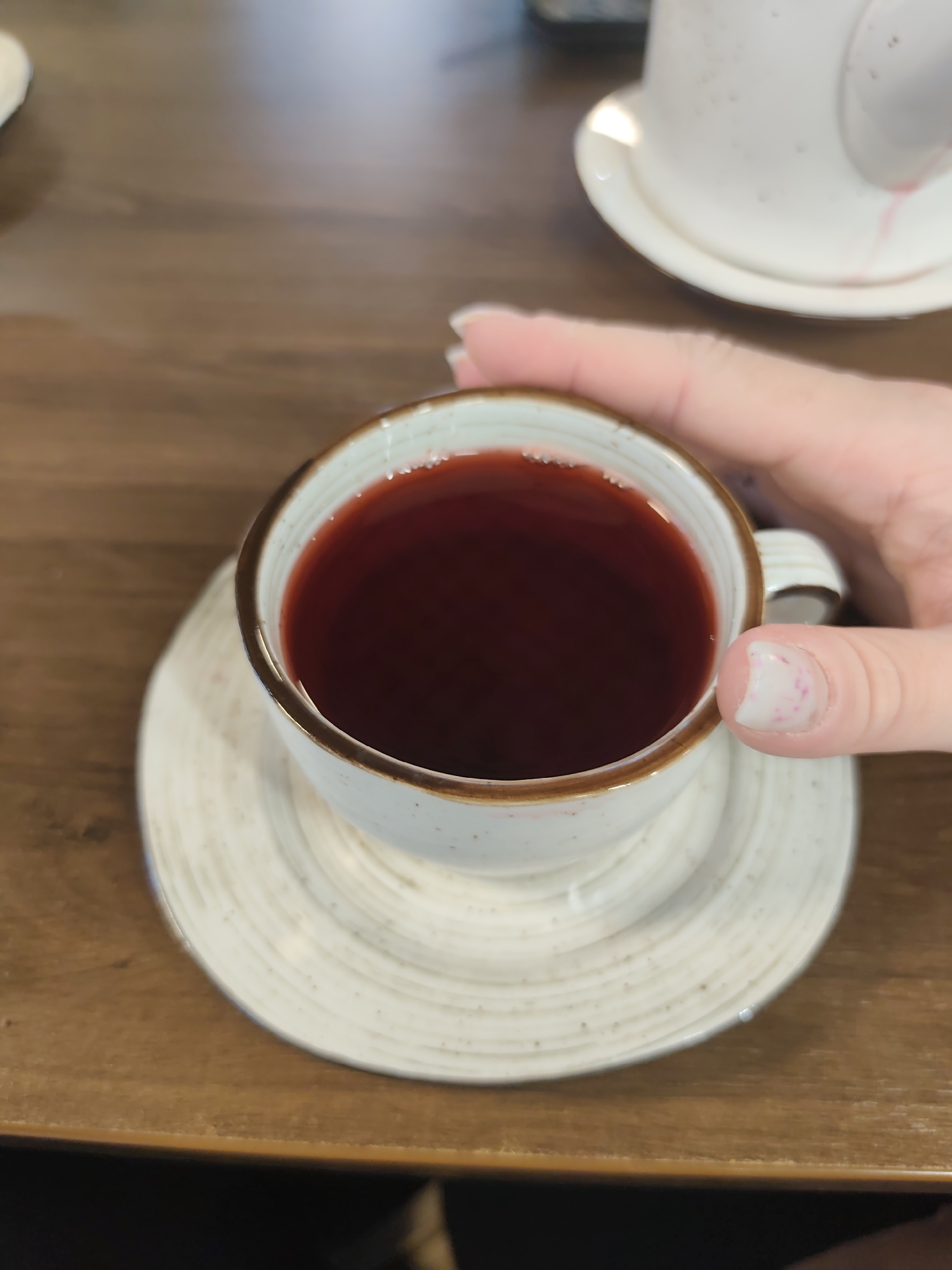 An overhead shot captures a person's hand holding a cup of dark red liquid, likely a hot beverage, on a wooden table in Chaikovsky, Russia. The cup is off-white with a speckled texture and a brown rim, resting on a matching saucer with concentric ridges. The liquid inside the cup is a deep, rich red, suggesting a berry tea or fruit drink.
The person's thumb and part of their fingers are visible, gently gripping the cup's handle. The thumbnail shows natural growth with a crescent-shaped white area and some faint, scattered pinkish discoloration, possibly from faded polish or contact with the drink.
In the background, out of focus, another white ceramic cup and saucer can be partially seen, suggesting a cafe or dining setting. The wooden table surface is light brown, providing a warm, natural backdrop for the scene. No text is visible on any of the objects. The image conveys a calm, possibly leisurely moment centered around enjoying a drink.