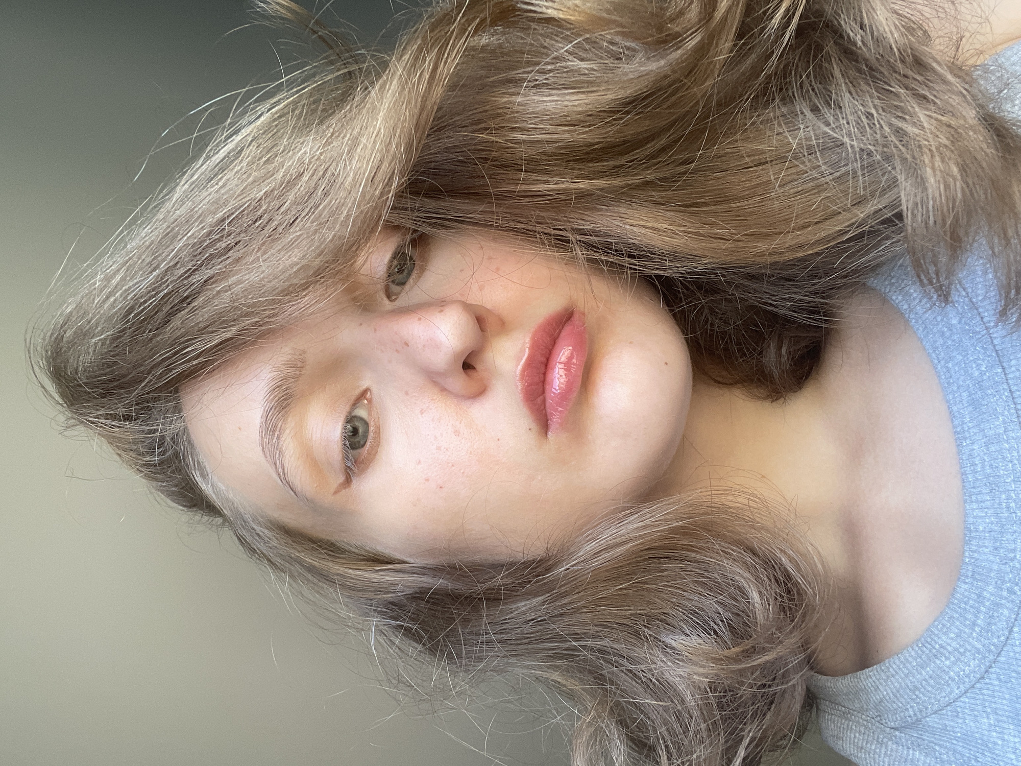 A close-up portrait shows a young person with light-colored hair and fair skin. The hair is a mix of blonde and light brown shades with wavy texture. The person has visible freckles on their nose and cheeks. Their lips are coated with a glossy pink lip product. One eye is visible, appearing to be green or hazel in color. The person is wearing a light-colored, possibly grey, garment. The background is a muted, neutral tone. The scene's location and connection to Подшивалово, Russia, are unknown.