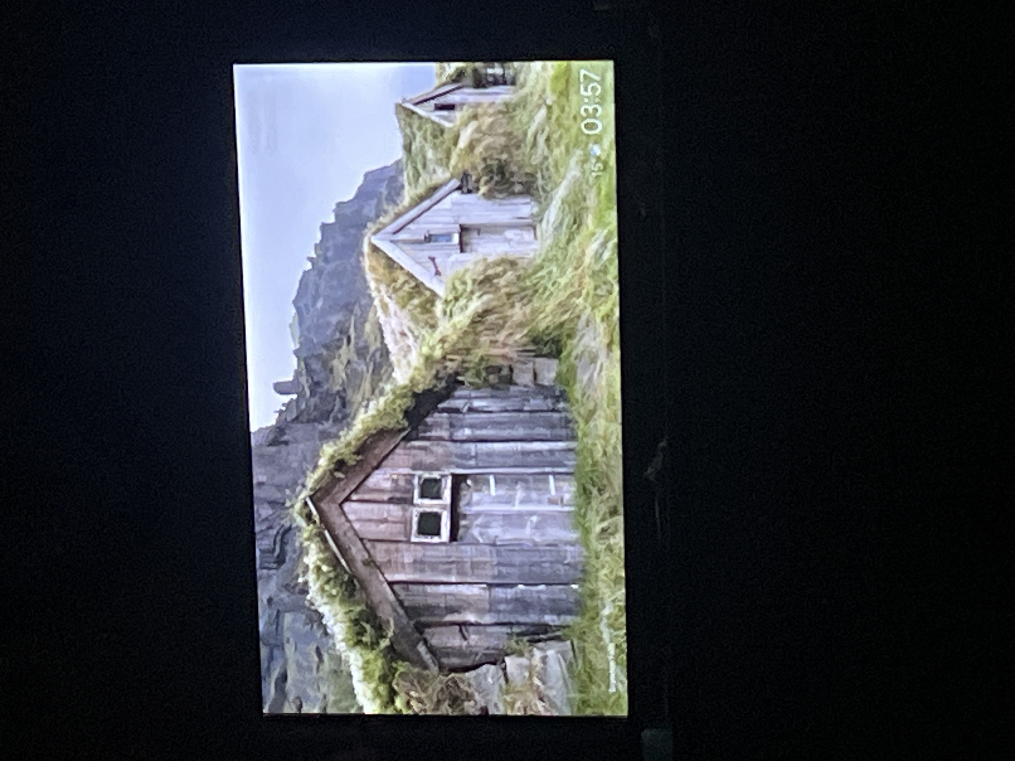 The image shows a scenic view of a cluster of small, traditional wooden huts with grass roofs situated on a grassy hillside. The huts have a rustic appearance, with weathered wooden planks forming their walls and simple windows. The surrounding environment is characterized by rugged, rocky terrain in the background and lush, green grass covering the ground and the roofs of the huts, suggesting a natural, possibly remote location. There is a visible digital clock in the lower right corner of the image displaying "03:57". The overall impression is one of a peaceful, natural landscape with historical or cultural significance. There is no indication of the city of Zarudyntsi, Ukraine in the image.
