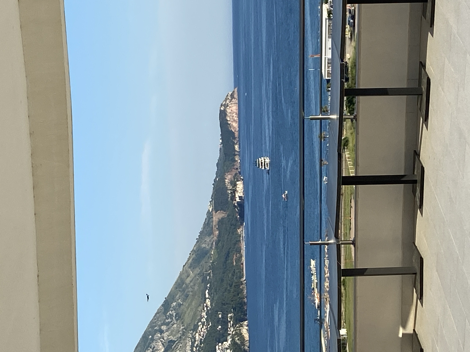 View of the Bay of Kotor, Montenegro, from a balcony. A mountain extends down the right side of the image, with a few small buildings visible on it. A large body of water extends down the center of the image, with a few sailboats and ships in the water. The sky is clear and blue. A bird is flying in the sky on the left. The balcony's glass and metal railing are on the right.
