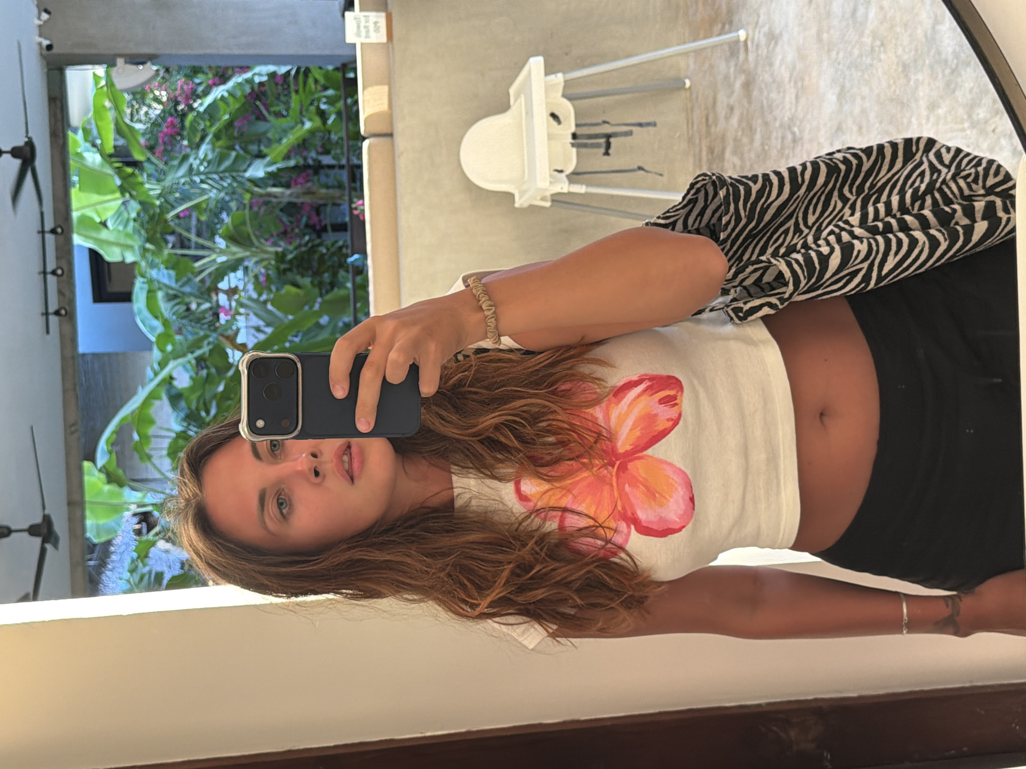 A young woman is captured in a mirror selfie, standing indoors in what appears to be a modern setting such as a cafe, hotel, or resort. She has long, wavy light brown hair, blue-green eyes, and is looking directly at the camera while holding a dark-colored smartphone, likely an iPhone, to take the picture. She is wearing a white crop top with a vibrant pink and orange plumeria or hibiscus-like floral design on the front, black shorts, and a light beige scrunchie on her right wrist. Her midriff is visible.
The reflection in the mirror reveals a lush tropical outdoor environment through a window or opening behind her. This outdoor scene is filled with large green leaves, possibly banana plants, and clusters of small pink or purple flowers, consistent with a tropical location like General Luna, Philippines. Also reflected in the room are a white, modern high chair with silver legs, a black ceiling fan mounted on the ceiling, and a black and white zebra-print bag or piece of fabric draped over something to her right. A small, white rectangular sign with unreadable text is reflected in the upper right corner of the mirror.
The natural light suggests it is daytime, illuminating the outdoor scene. The indoor space has a concrete or minimalist aesthetic. The overall impression is a casual moment captured in a pleasant, tropical setting.