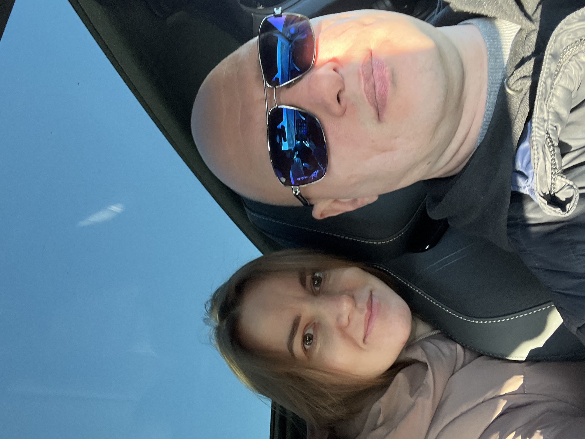 The image shows two people in a car. A bald man wearing sunglasses is in the background, and a woman with light brown hair is in the foreground. The environment visible outside the car appears to be a clear, blue sky. There are no indications of the location being Malosofiivka, Ukraine. The scene suggests a casual selfie taken inside a vehicle.