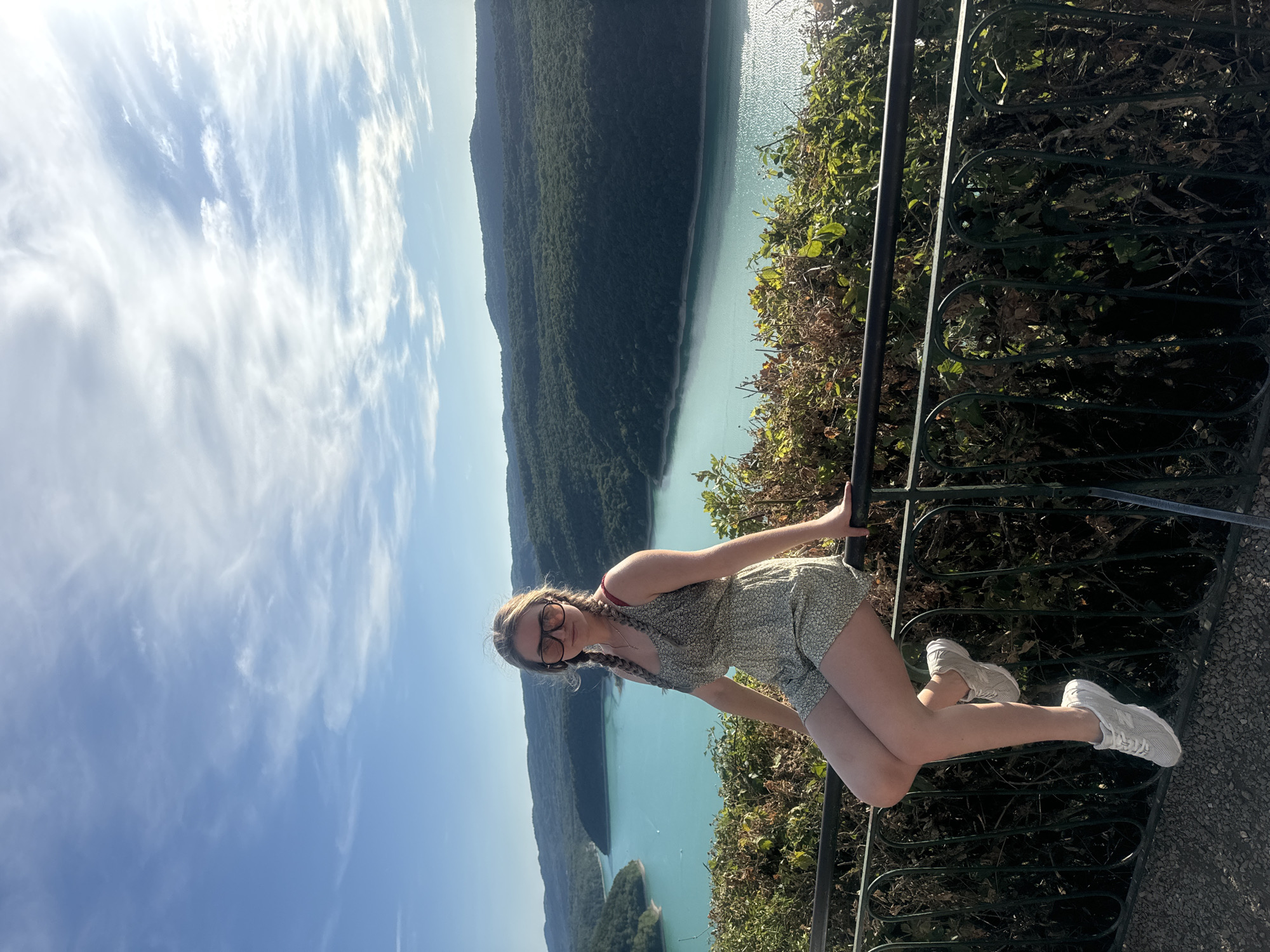A young Caucasian woman with blonde hair in braids and sunglasses is seated on a dark metal railing. She is wearing a floral print romper and white sneakers. She is looking at the camera. Behind her, a body of turquoise water, likely a lake or river, is surrounded by lush green, tree-covered hills. The sky is blue with scattered white clouds. The scene appears to be an overlook, possibly in Moirans-en-Montagne, France, known for its scenic beauty and natural landscapes.