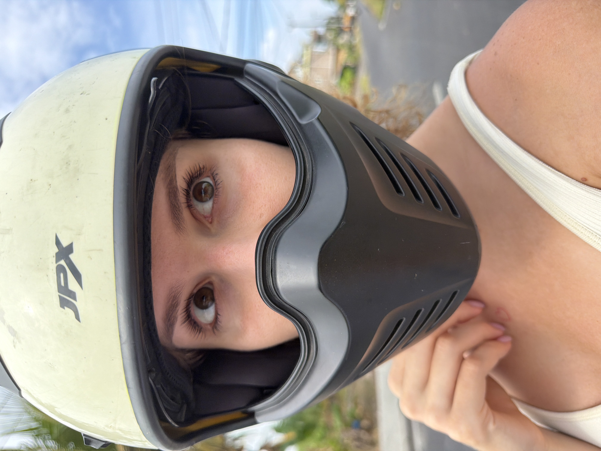 A medium shot from the chest up features a young woman wearing a light yellow motorcycle helmet with black trim and a black face mask. Her brown eyes are wide open and looking slightly upward and to her right. The helmet has the "JPX" logo in black on its side. She is wearing a white tank top. Her left hand is raised to her neck, with light pink nail polish visible. The background is slightly blurred, showing a paved road, some greenery, and buildings, indicative of an outdoor setting in a city. The sky is visible, with scattered clouds. The scene is likely in Mengwi, Indonesia, given the vegetation and architecture.