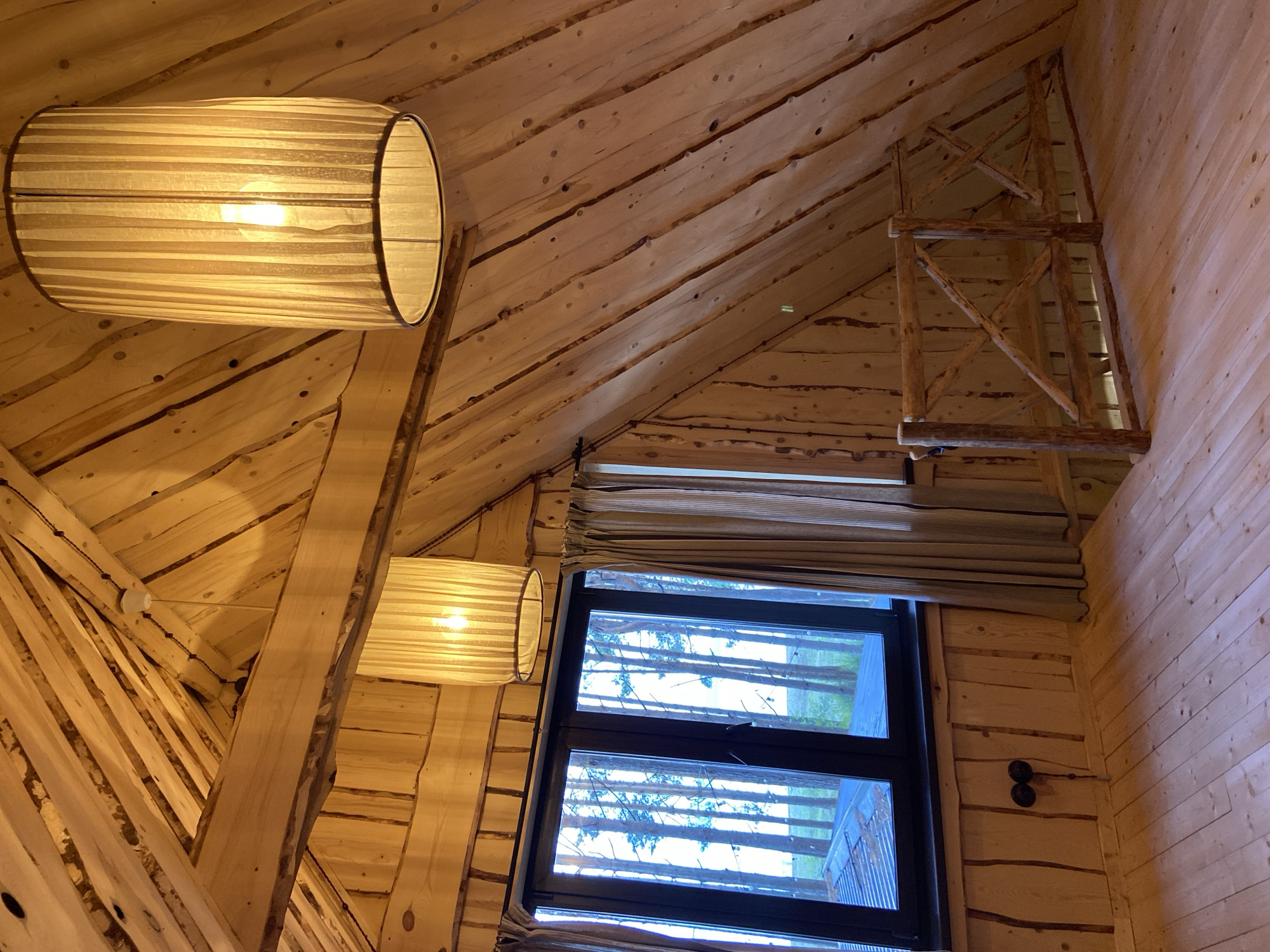 The image shows the interior of a wooden room with a sloped ceiling and walls, likely a loft or attic space, in a building located in Забегаево, Russia. The room features exposed wooden beams and paneling, giving it a rustic aesthetic. Two large, cylindrical pendant lamps with textured fabric shades are hanging from the ceiling, illuminating the space. A large, dark-framed window is on the left wall, offering a view of tall pine trees and a bright sky, suggesting an outdoor environment. Light brown curtains hang on either side of the window. In the foreground and to the right, there is a wooden railing with a criss-cross design, indicating a potential balcony or a separation of space. The floor is also made of light-colored wooden planks. Two circular electrical outlets are visible on the wooden wall below the window. The overall impression is of a cozy, unfinished wooden interior.