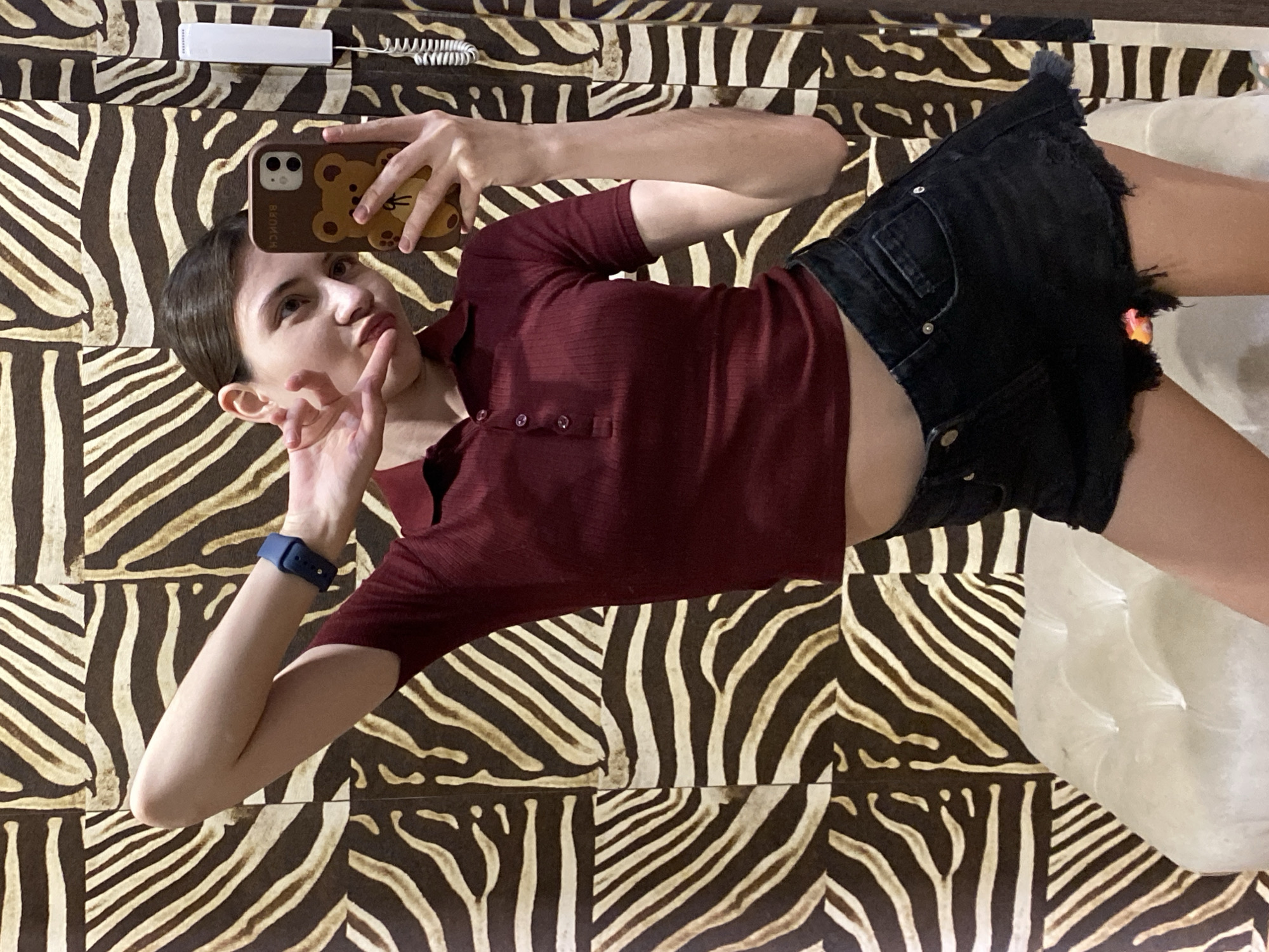 A person is taking a selfie using a smartphone with a brown bear case while lying on a bed with zebra-patterned sheets. The person is wearing a maroon-colored, ribbed, button-up shirt, a blue wrist wearable device, and black denim shorts. A white landline phone is in the upper left corner. A white pillow is in the lower right corner. The location appears to be indoors, but there is no indication of it being in Votkinsk, Russia.