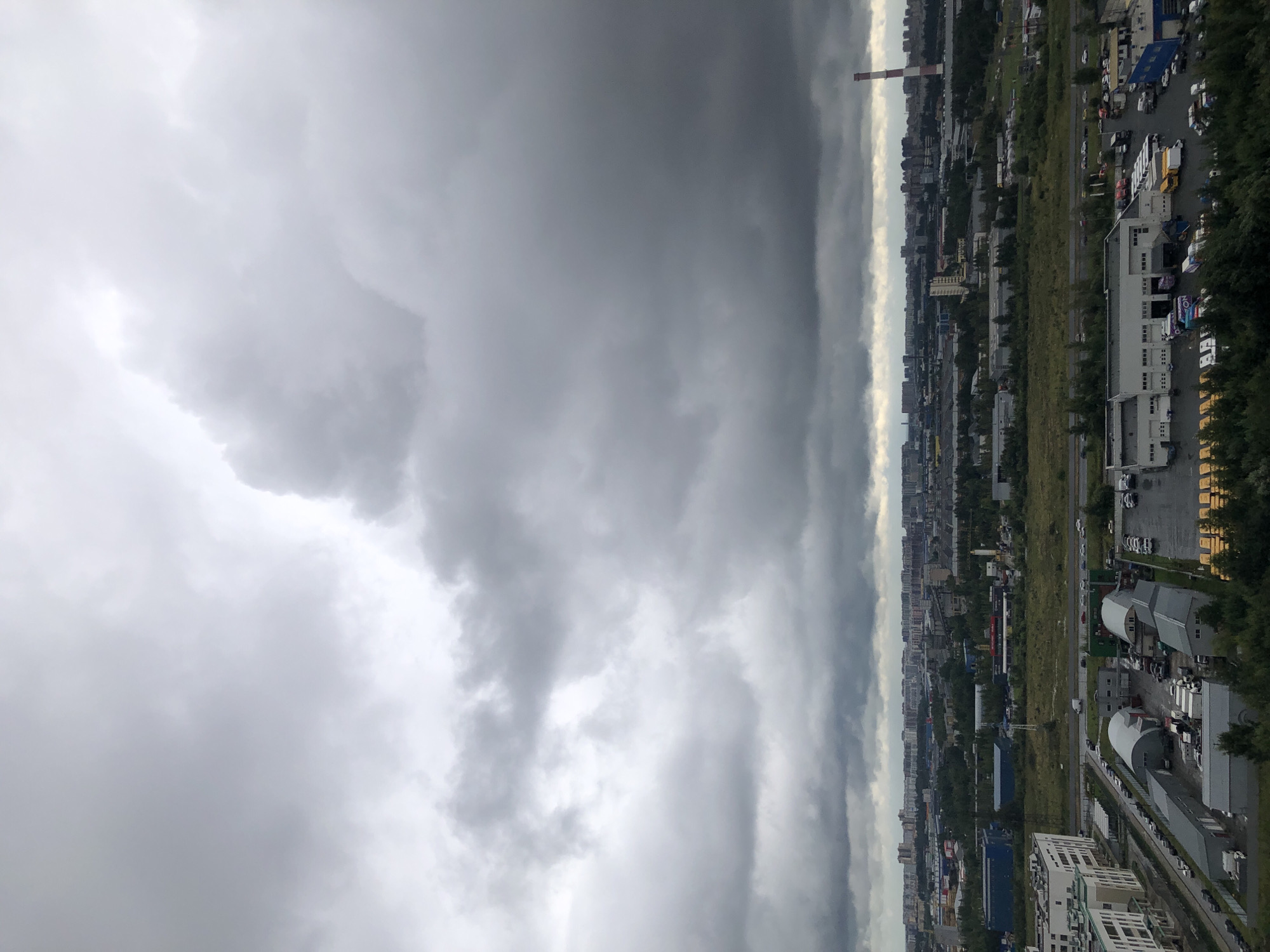 The image displays an aerial view of a city under a heavily overcast sky. The foreground features a modern residential building on the left, followed by a complex of industrial warehouses and parking lots filled with vehicles, including trucks and vans. A road with several cars is visible in the middle ground, separating the industrial area from a large expanse of greenery and scattered industrial buildings. In the background, a dense cityscape with numerous buildings stretches across the horizon, interrupted by a tall, red-and-white striped chimney on the right. The overall atmosphere is somber due to the dark, textured clouds dominating the upper half of the frame, suggesting an impending storm or inclement weather.