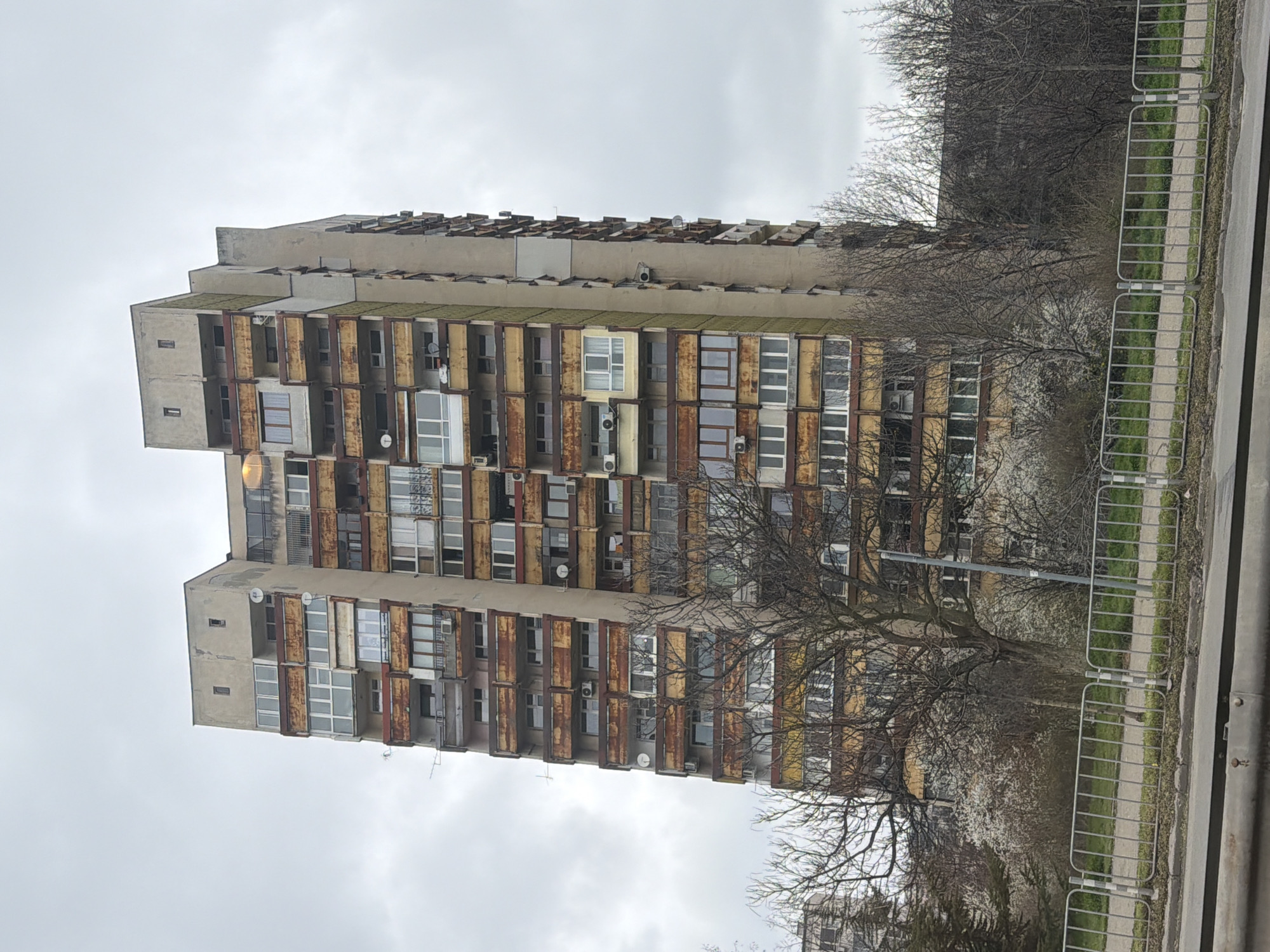 A medium shot depicts a Soviet-era residential building, likely in Sofia, Bulgaria, evidenced by architectural style. The building is tall and characterized by a prefabricated concrete structure. It is divided into multiple stories, each containing apartments. Each apartment has a recessed balcony. The building's facade shows signs of age and weathering, including discoloration and rust. Satellite dishes are visible on some balconies, indicating modern occupation and use. In the foreground, a metal fence runs along a sidewalk or street. Trees are bare or in early bloom, with branches obscuring parts of the building. Overcast skies are present.