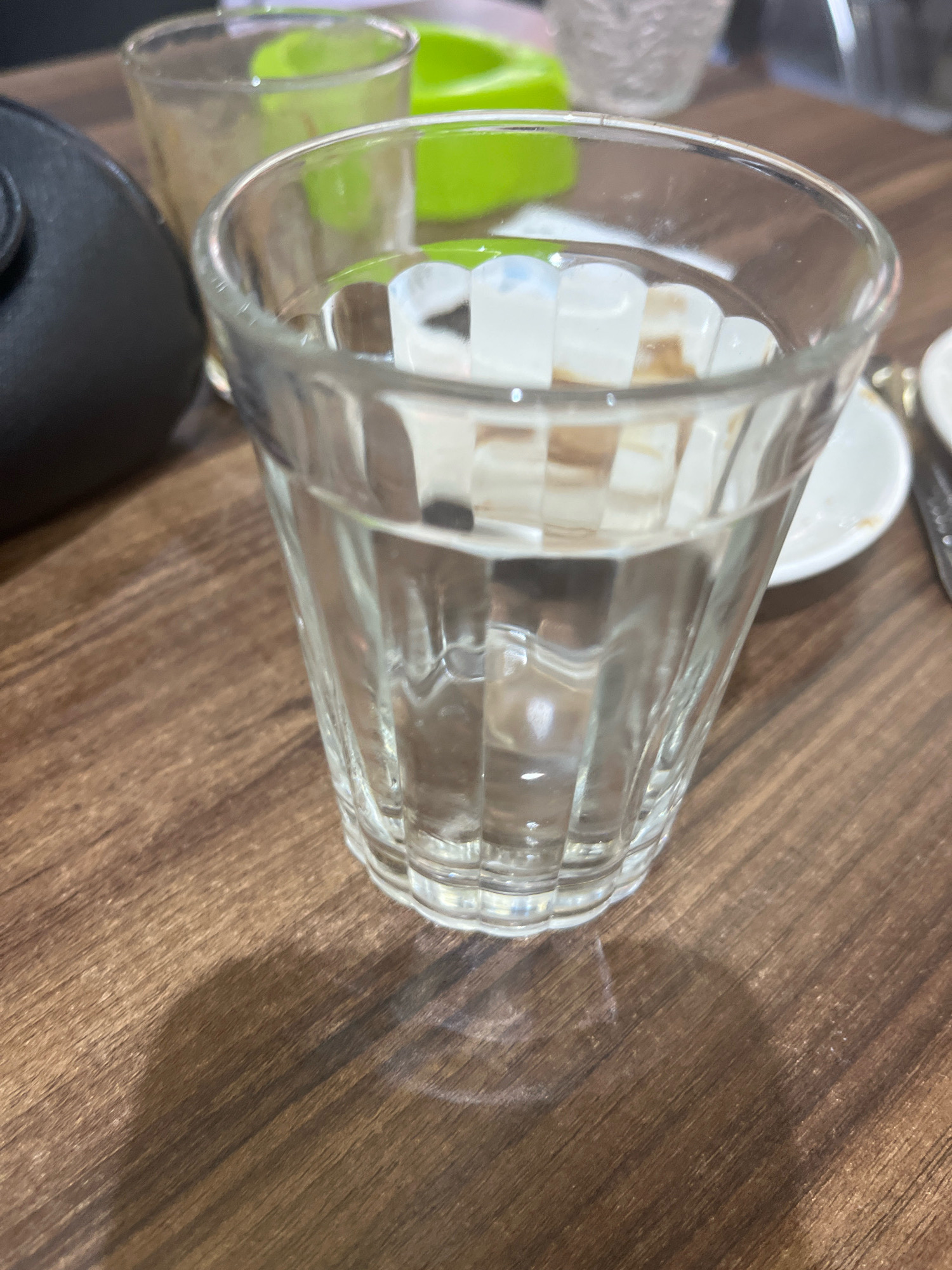 A close-up shot shows a glass of water on a wooden table. The glass is clear and has vertical ribbed details. There are ice cubes visible inside the glass. A black object, possibly a purse, is partially visible in the upper left corner of the frame. In the background, out of focus, are other glasses and a green object. The table has a brown, grainy texture. The lighting creates a shadow of the glass on the table. The image appears to be taken in Azilal, Morocco.