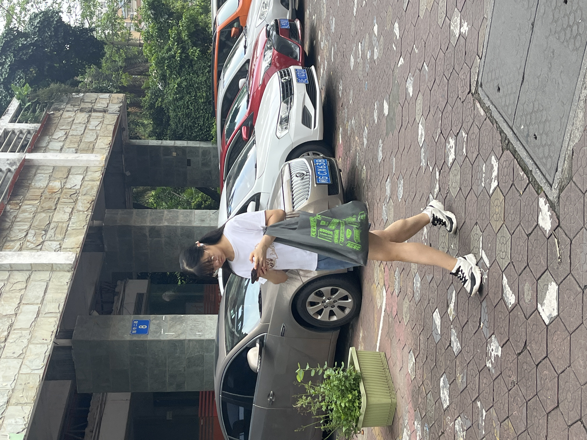 A young East Asian female, wearing glasses, a white t-shirt, blue shorts, and black and white sneakers, walks across a brick-patterned sidewalk while looking at her phone. She carries a large grey tote bag with green lettering over her right shoulder. To her left, a row of parked cars are visible, including a silver sedan, a white sedan, a red sedan, and an orange SUV. In the background, a building with a stone facade and pillars is present, along with lush green trees and bushes. A blue sign with white Chinese characters and the number "8" is affixed to one of the pillars. The scene appears to be an outdoor parking area in Chancheng District, China.