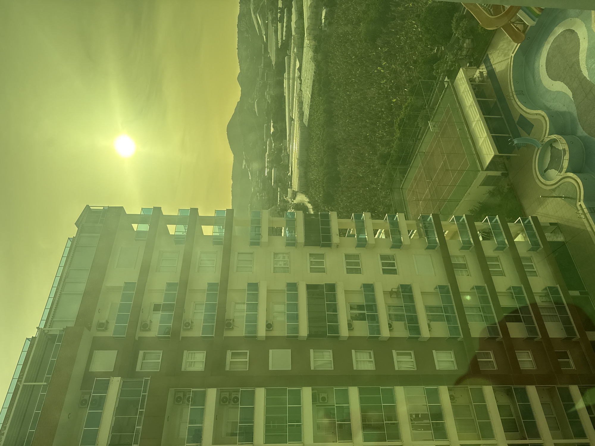 The image shows an exterior view of a tall building with many windows. The building is located in a city in an unknown location. The environment includes the bright sun in the sky, a distant mountain, some greenery, a tennis court, and a swimming pool. There is no people in the image.