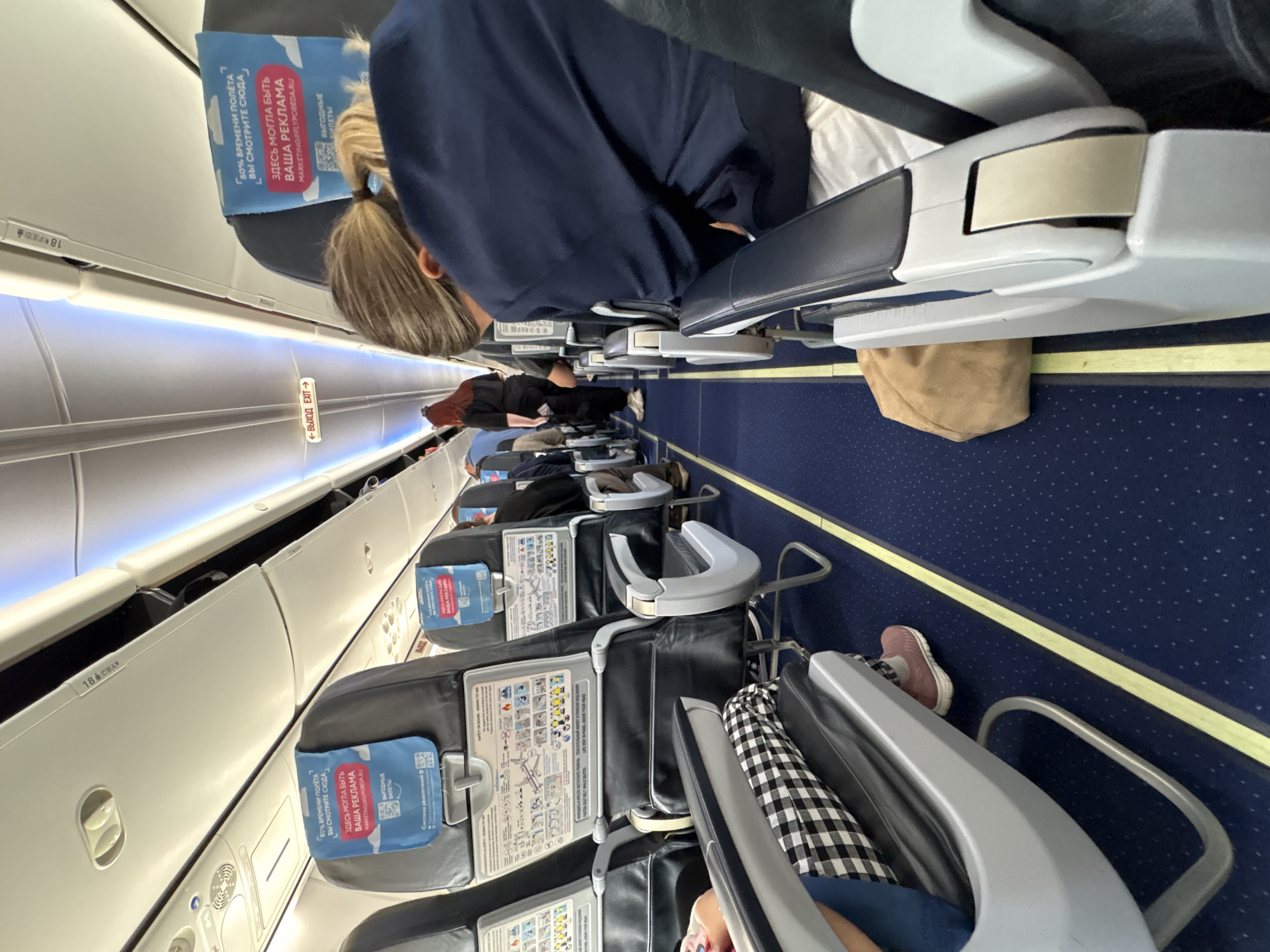 The image depicts the interior of an airplane cabin, viewed from a passenger's perspective, facing forward. The plane has rows of seats, each with a headrest and safety information card. Passengers are seated, some resting or occupying themselves, with legs and belongings visible in the aisle. Overhead compartments run along the top of the cabin, illuminated by interior lights. The carpet is patterned in blue, and advertising material is displayed on the seats.