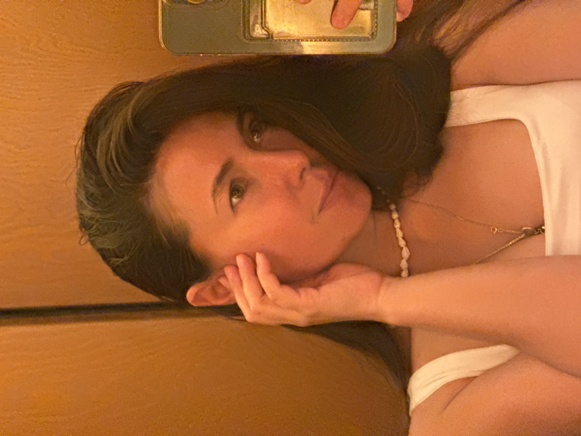 A woman with dark hair is lying down, resting her head on her hand, while taking a selfie. She is wearing a white top and two necklaces, one with pearls. A smartphone is partially visible in the top of the frame. The background is a blurred warm-toned wall. There is no indication of the location being Kazan, Russia, or any other city.