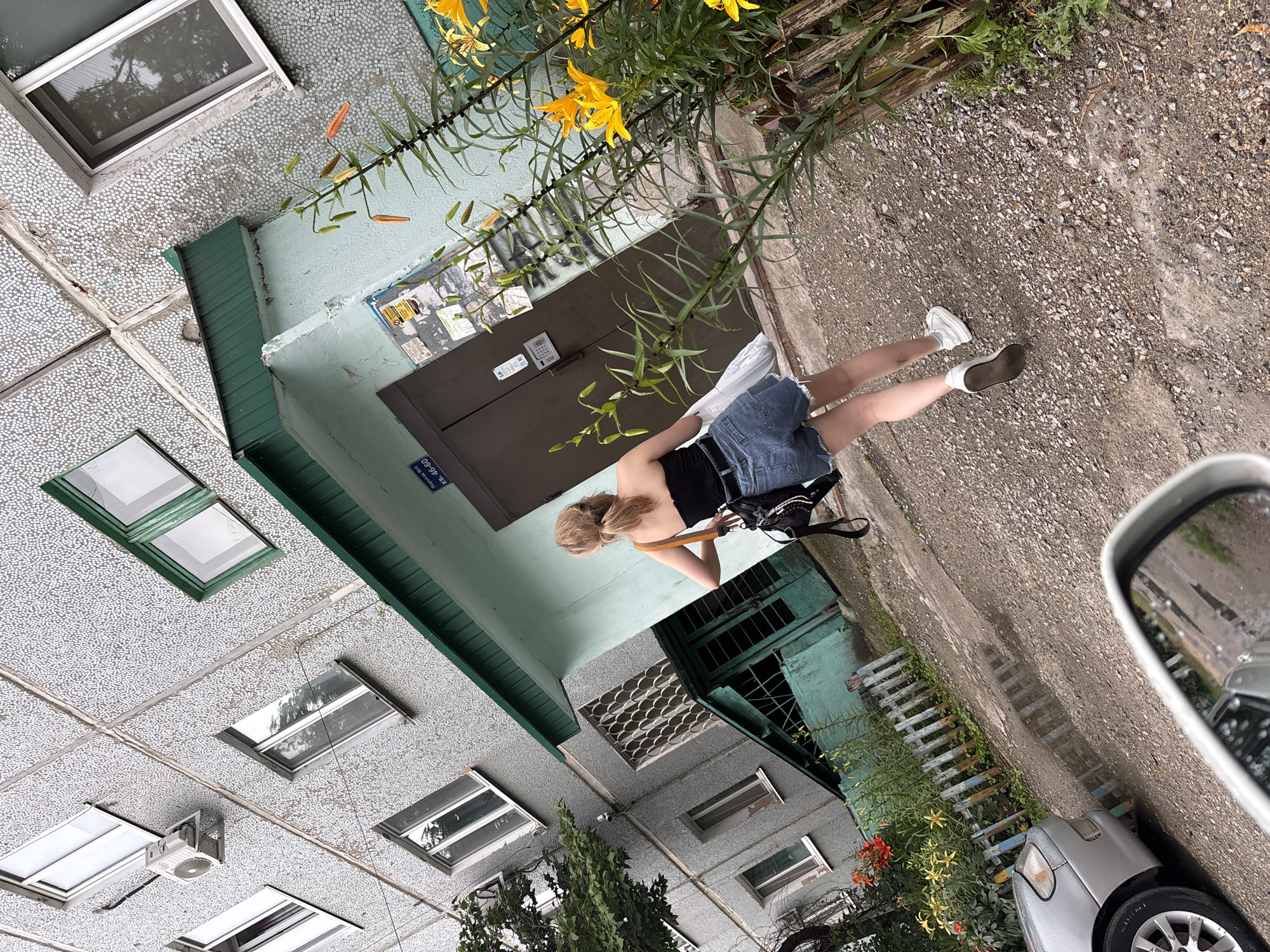 A young woman with blonde hair pulled back in a ponytail walks away from the camera towards a building entrance. She is wearing a black strapless top, denim shorts, and white sneakers. She carries a white plastic bag in her right hand and a black bag over her left shoulder.
The building is a multi-story apartment building with a gray, textured facade. The entrance has a light blue facade with a dark brown door and a green overhang. A sign above the door reads "Подъезд №4" (Entrance No. 4) and "ул. 40-60" (40-60 Street). There are several windows visible on the building's facade, some with green trim.
In front of the building, there is a patch of ground covered with gravel and dirt. A small wooden fence runs along a flowerbed containing blooming orange and yellow lilies. To the left, a silver car is partially visible. Puddles of water are present on the ground, indicating recent rain. The scene appears to be in an urban residential area.
This image was taken in Blagoveshchensk, Russia. The street sign visible in the image, "ул. 40-60," refers to "40 Years of the Komsomol Street," which exists in Blagoveshchensk.