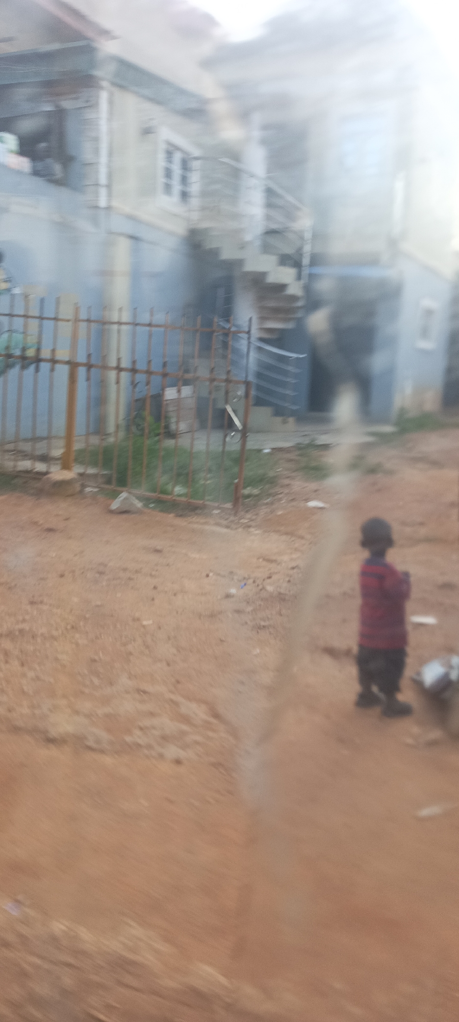 The image presents an outdoor residential scene in Bukuru, Nigeria, captured with a noticeable blur, possibly due to camera movement or being taken through a slightly obscured window.
The foreground and much of the midground are covered by reddish-brown, unpaved ground, indicative of dirt or sand. Stretching across the mid-left of the frame is a rusty, metal fence composed of vertical bars, providing a boundary to a small patch of green grass directly behind it. A light-colored stone or concrete object lies near the base of the fence.
To the right, a small child stands with their back turned to the camera. They are dressed in a red and black striped long-sleeved shirt and dark lower garments. A small, light-colored object is visible on the ground beside the child.
In the background, a multi-story building, painted in light blue or grey tones, occupies the space behind the fence. This structure features several white-framed windows and an external spiral staircase with white railings. Another individual is faintly visible through an open window on the upper left side of the building.
The scene is lit by natural daylight. The architectural style, the presence of unpaved ground, and the type of fence are consistent with residential settings in the specified location. No specific activity is clearly depicted, and there is no visible text in the image.